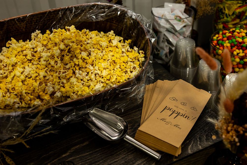 Large bowl of fresh buttery popcorn with a metal scoop beside paper popcorn bags on a snack bar table with candy in the background.
