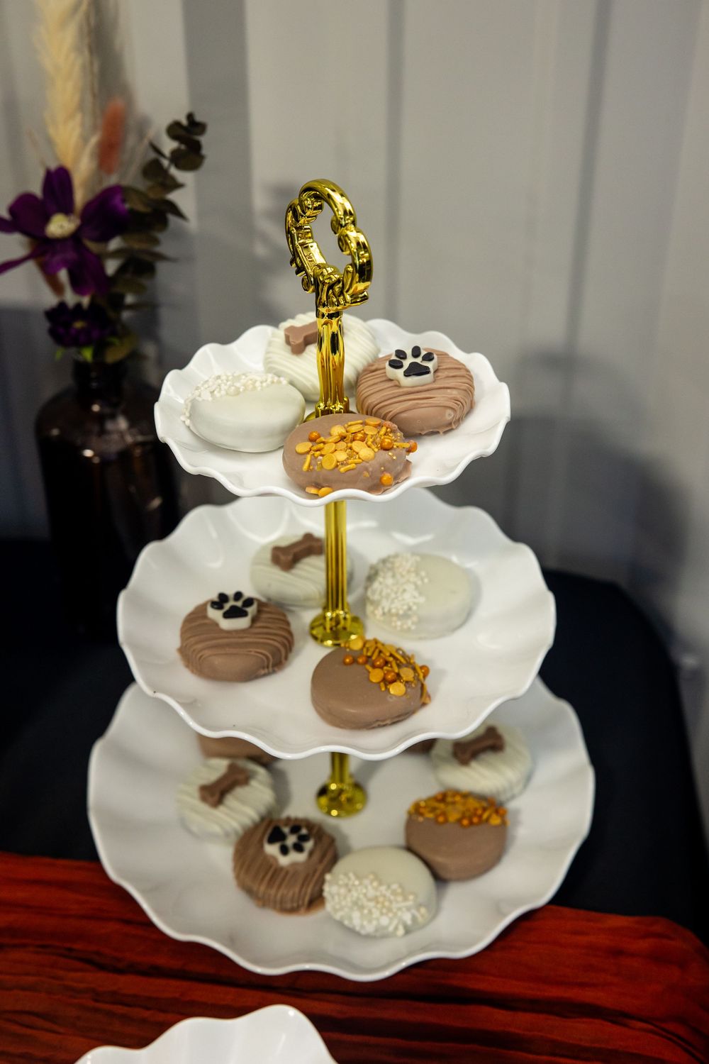 Three-tier white ruffled dessert stand with gold handle holding chocolate-covered sandwich cookies decorated with paw prints and sprinkles.