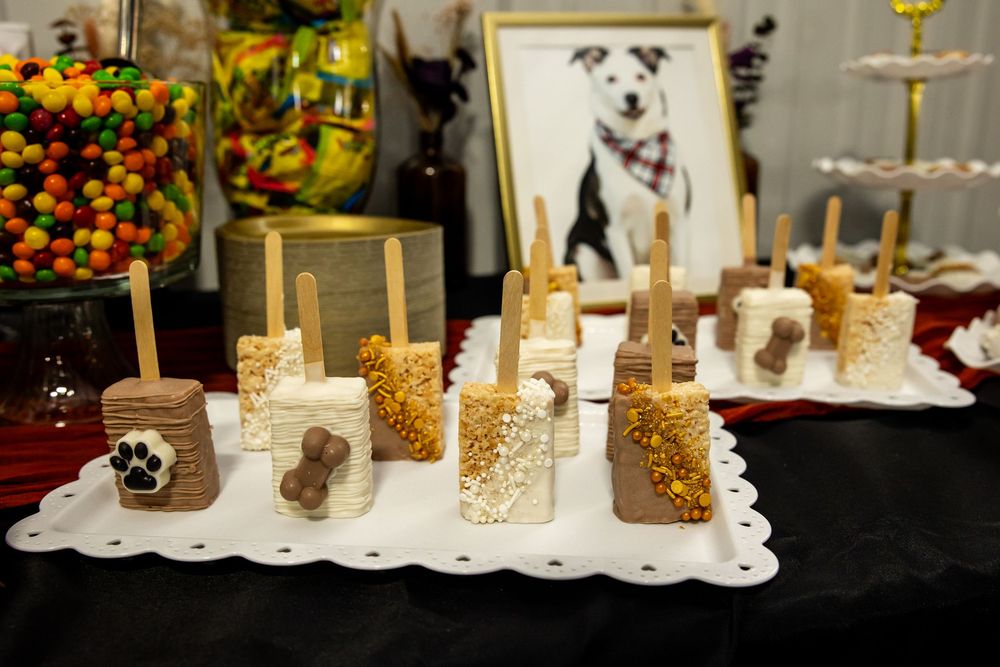 Dog-themed dessert table with paw print and bone decorated treats on sticks, a candy jar and a framed dog portrait in the background.
