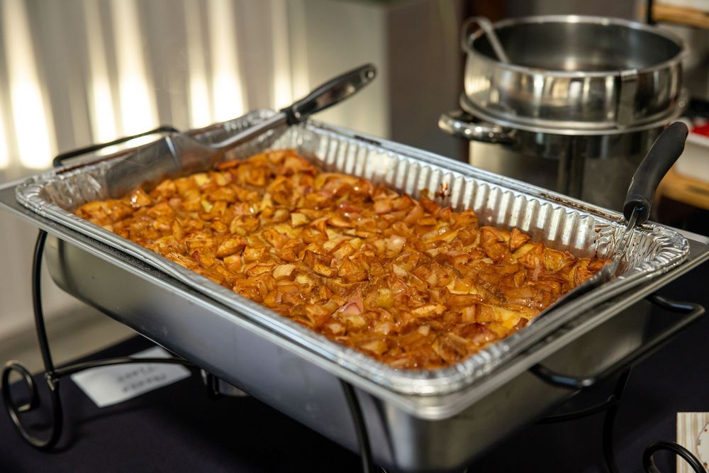 Foil-lined buffet chafing dish filled with baked cinnamon apple dessert, served on a metal warming stand next to a stainless steel pot.