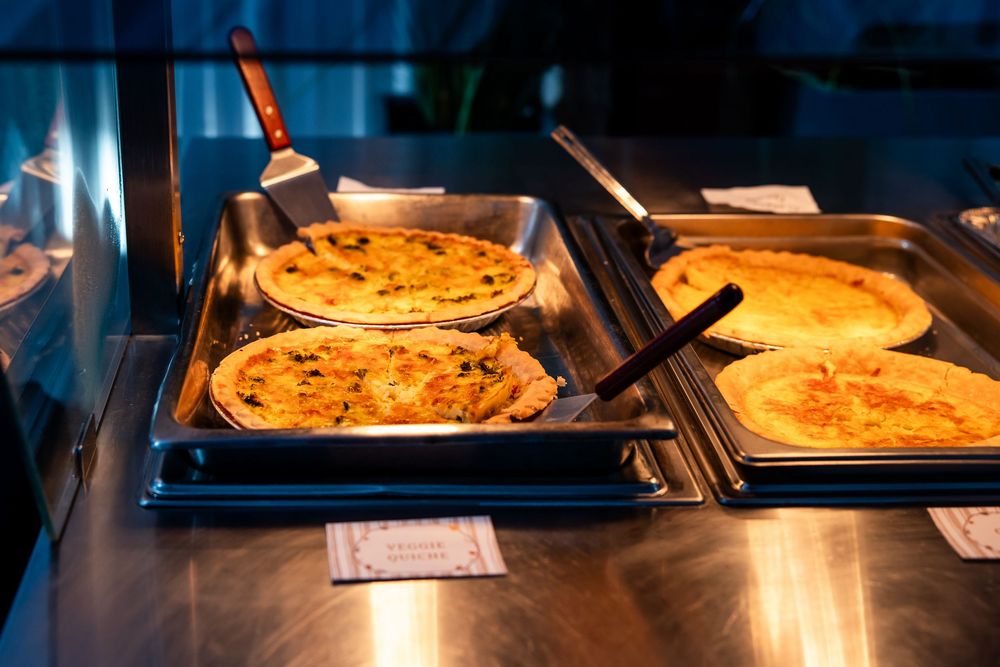 Four freshly baked quiches or savory pies sit in metal serving trays with spatulas on a cafeteria buffet line.