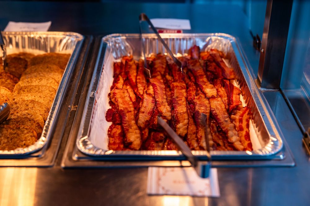 Metal serving trays of crispy bacon strips with tongs on a buffet line, next to a tray of golden hash browns.