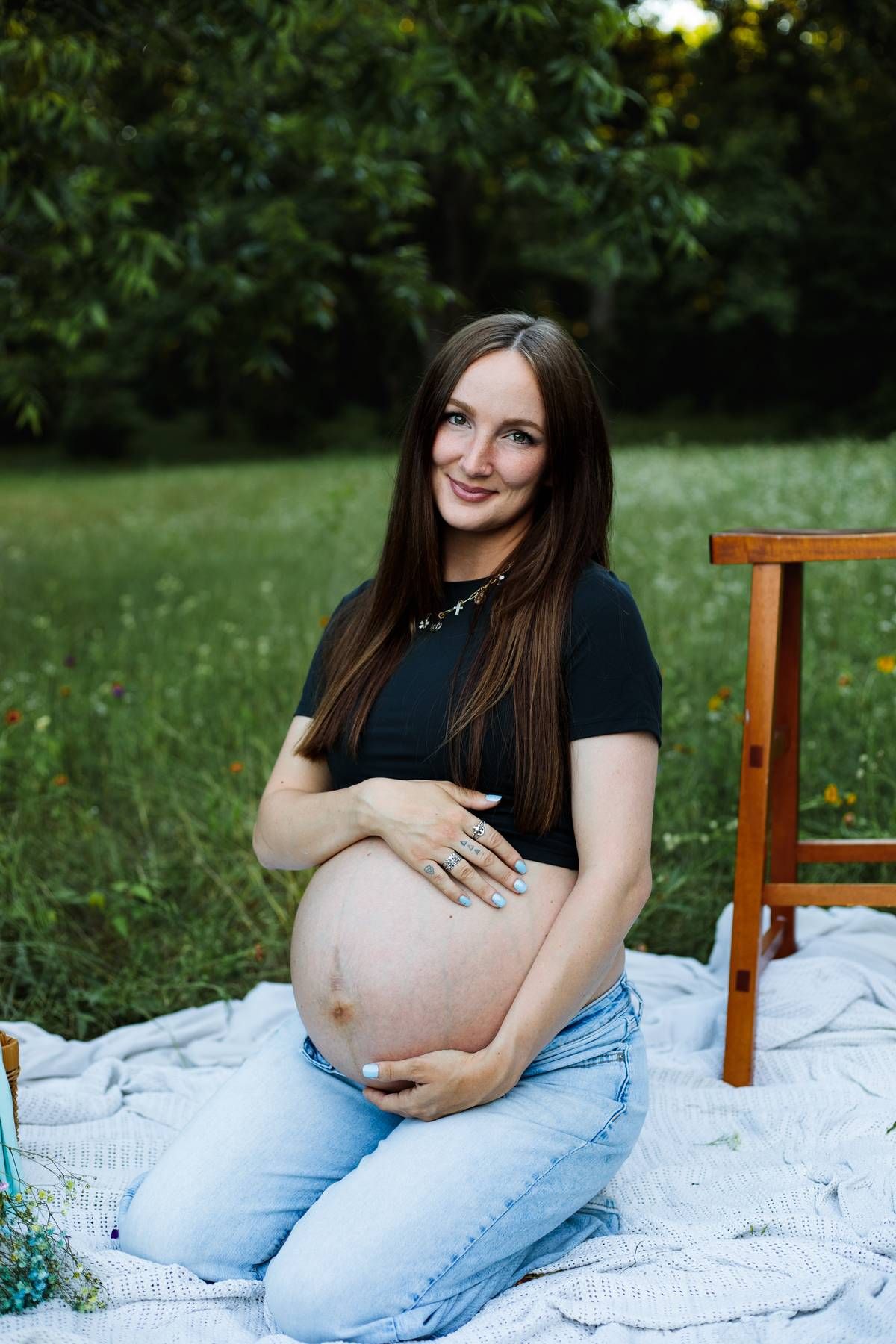 Pregnant woman in a black tee and jeans kneels on a blanket outdoors, cradling her bare baby bump and smiling.