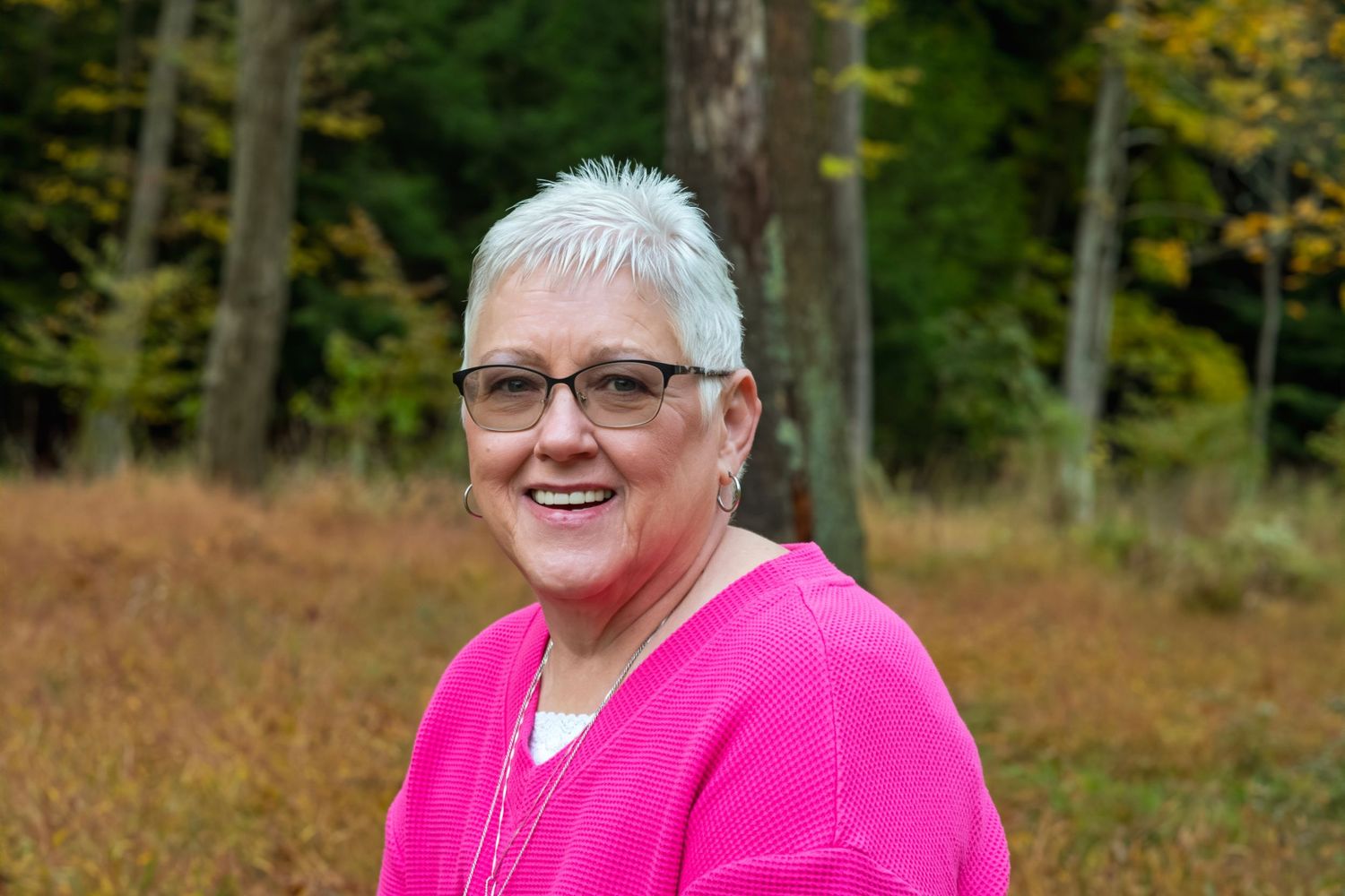 Smiling woman with short white hair and glasses wearing a pink sweater outdoors in a wooded autumn setting.