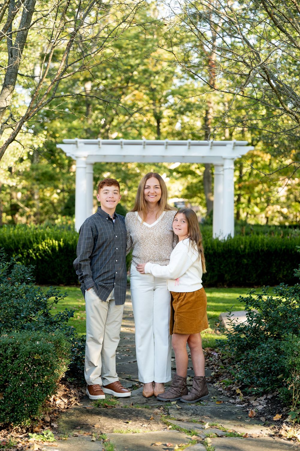 A woman and two children pose together in a lush garden with a white pergola backdrop.