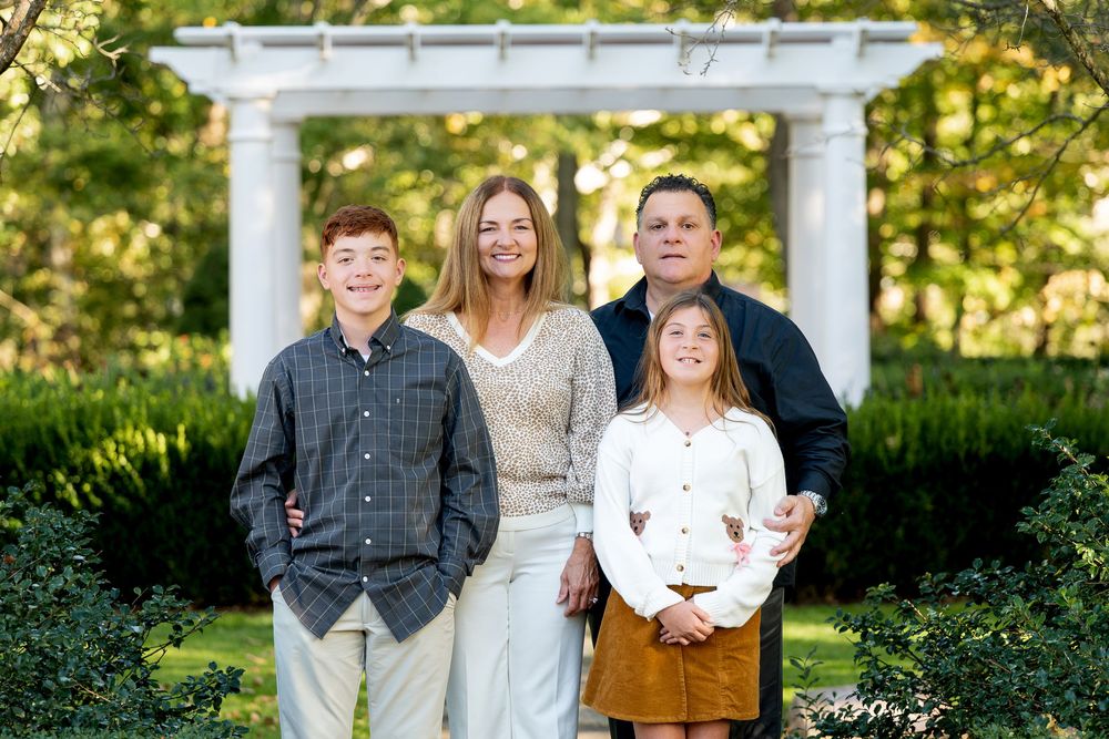 Family of four posing outdoors in a garden with white pergola and lush greenery in the background.