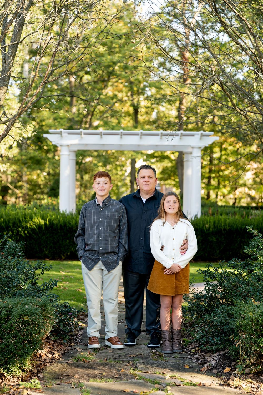Father poses with teenage son and daughter in an outdoor garden with white pergola.