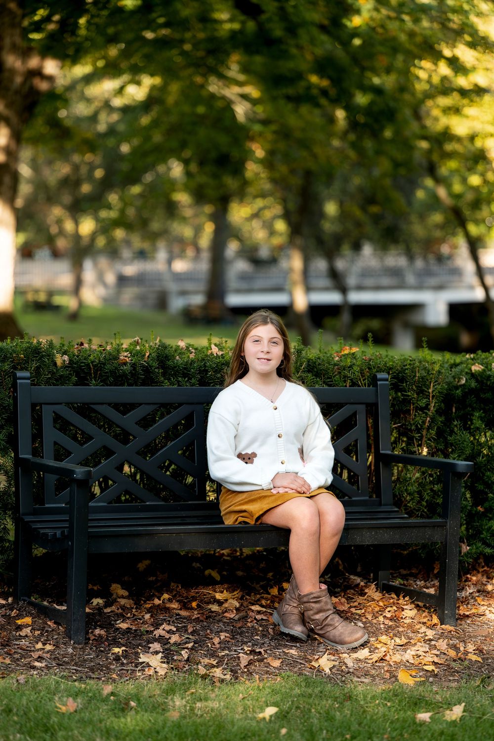 Teen girl in white cardigan and mustard skirt sits on black park bench surrounded by fall foliage.