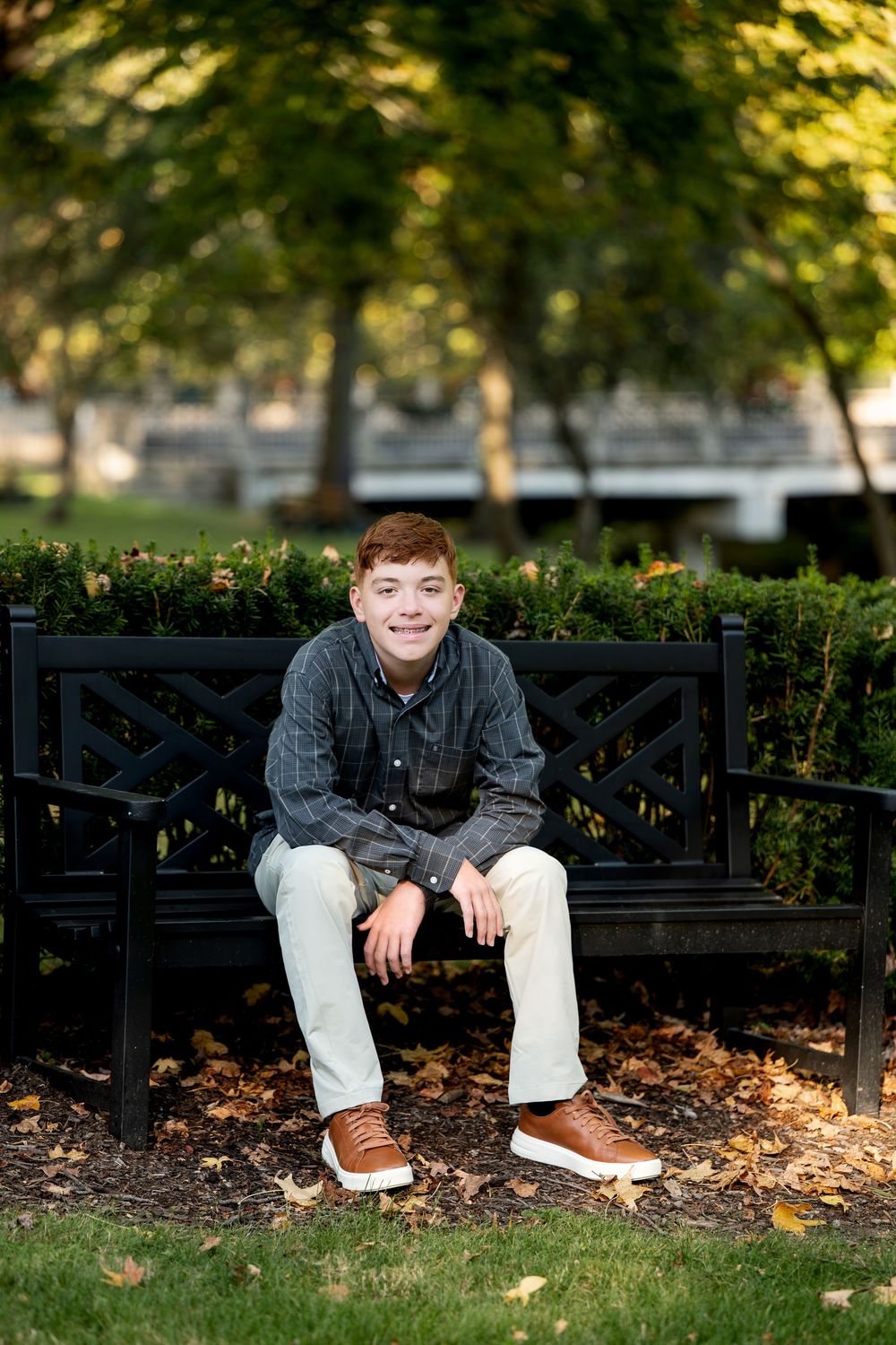 Smiling teenage boy sitting on black bench in park surrounded by autumn leaves.