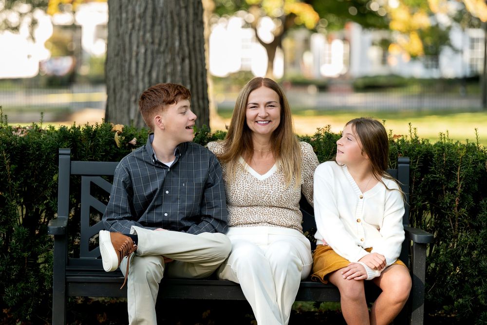 A woman sits on a park bench smiling with a teenage boy and girl in an outdoor setting.