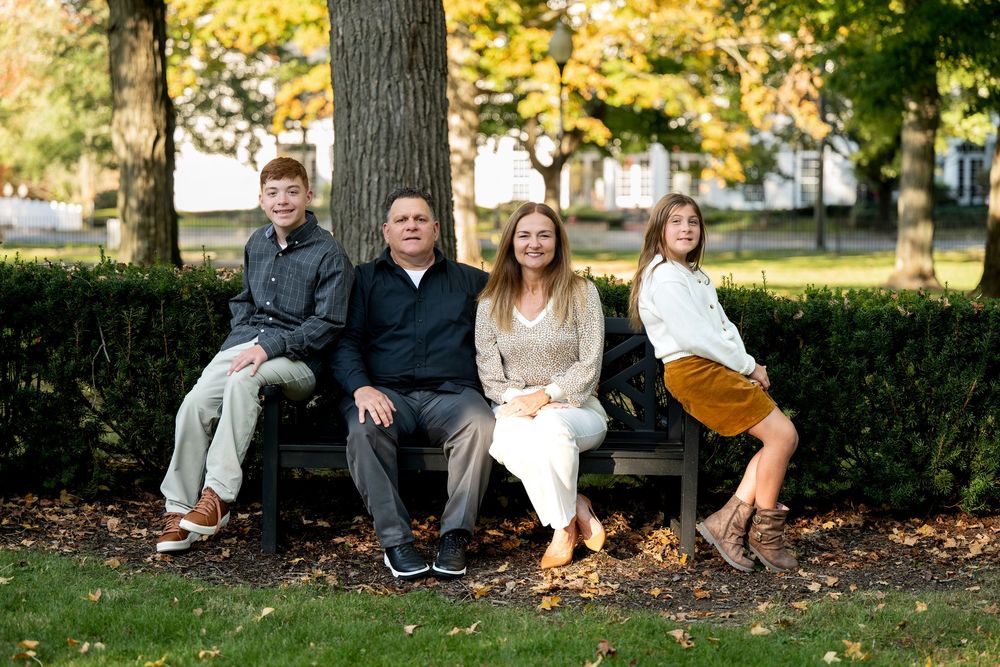 Family of four poses on park bench under large tree with colorful fall foliage.