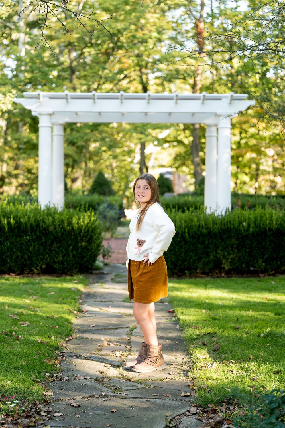 Smiling teenage girl in white sweater and tan skirt posing on a stone path with a white pergola in a green garden.