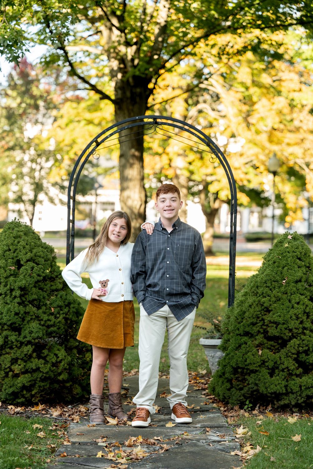 Teen boy and girl stand together under black garden arch surrounded by autumn landscaping.