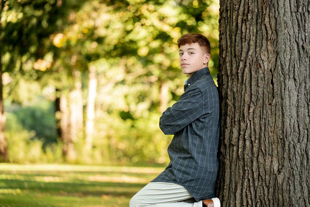 A teenage boy with red hair leans against a tree trunk with arms crossed, looking back in a green park.