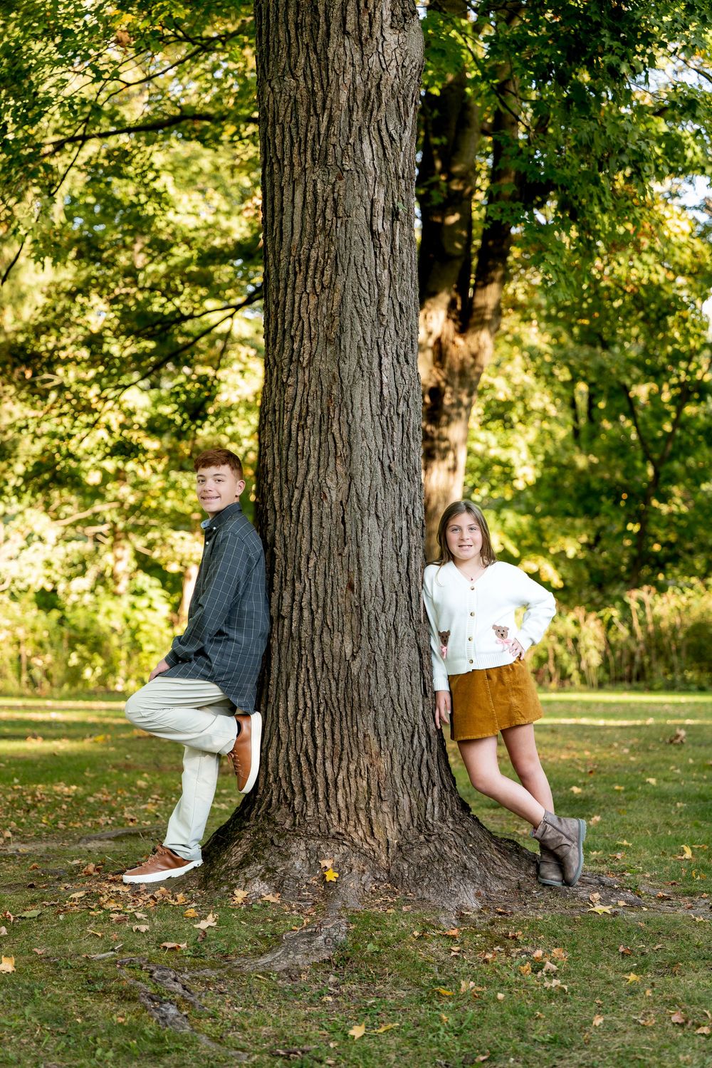 A teenage boy and girl pose on opposite sides of a large tree trunk in a lush green park setting.