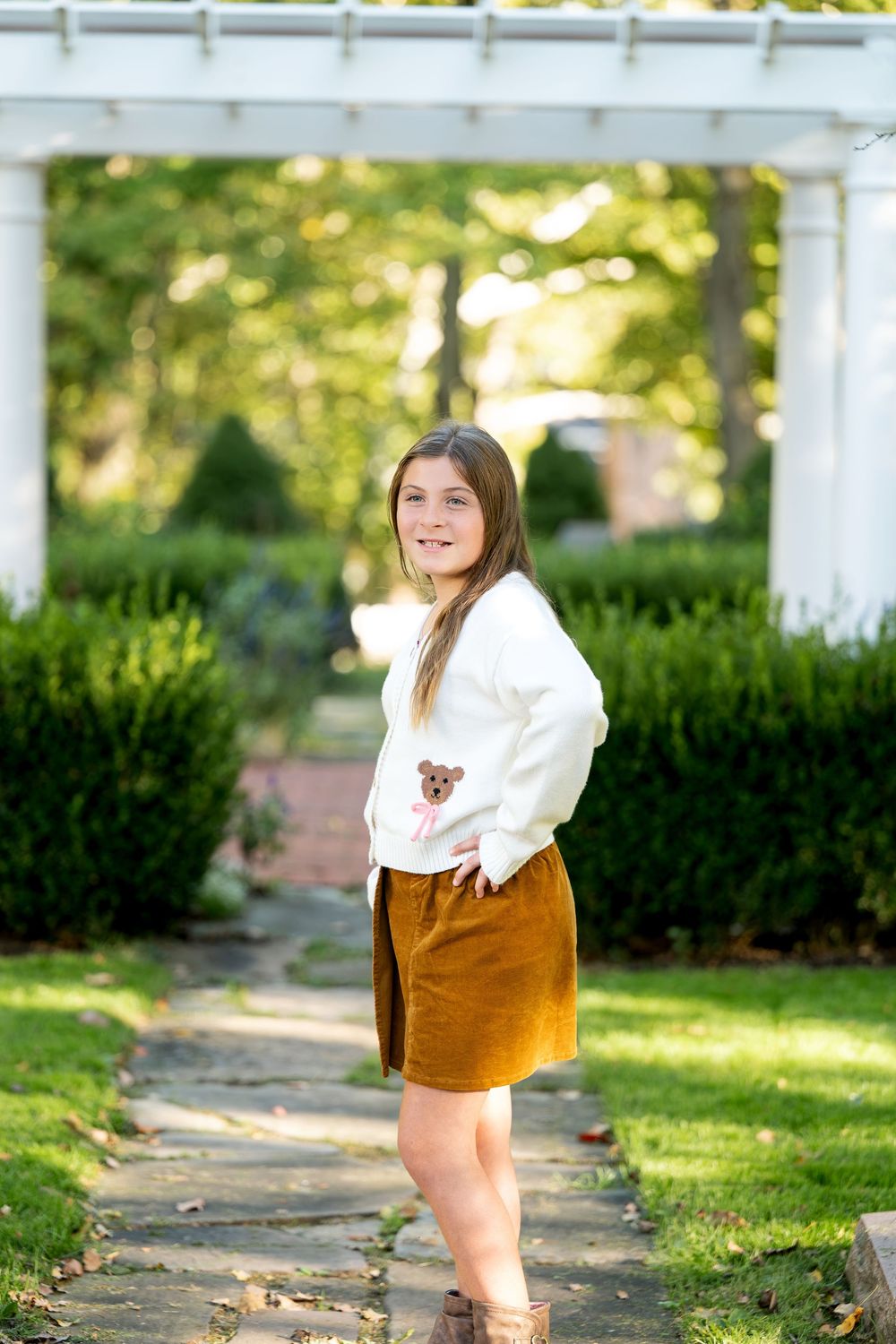 Young girl in white sweater and mustard skirt poses on garden path with white pergola and green hedges.