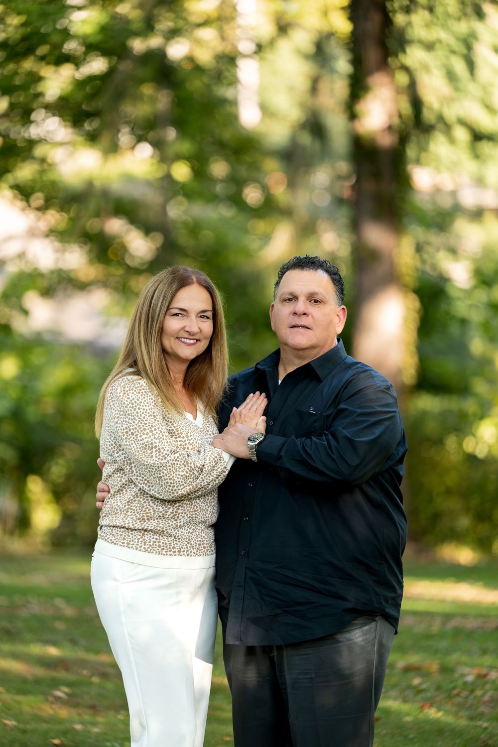 Happy couple embracing outdoors in a lush green park, woman in white pants and floral top, man in black shirt.