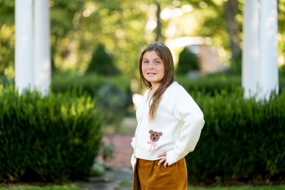 Young girl in white bear sweater and gold pants poses outdoors near white columns and green hedges.
