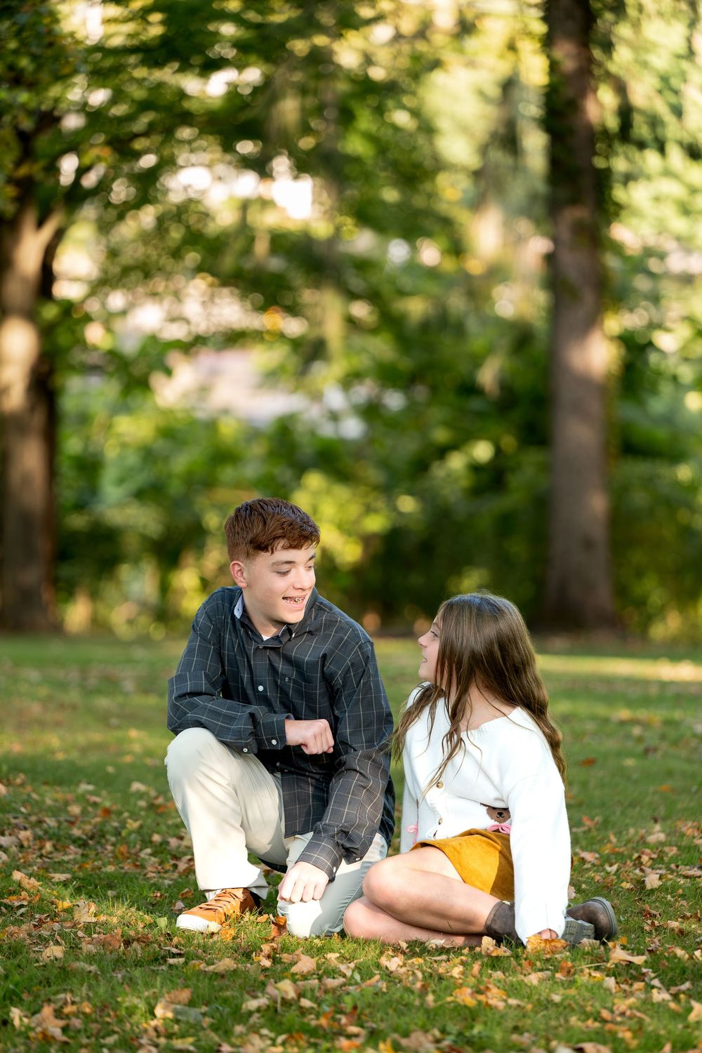 A teenage boy crouches beside a girl sitting on grass covered with fallen leaves in a park.