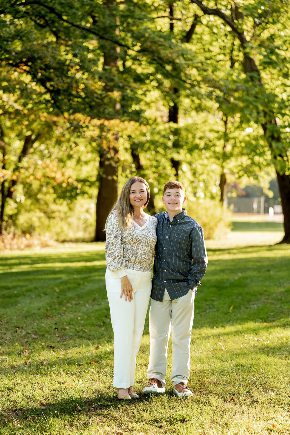 A woman and teenage boy smile together outdoors in a sunny park surrounded by lush green trees.