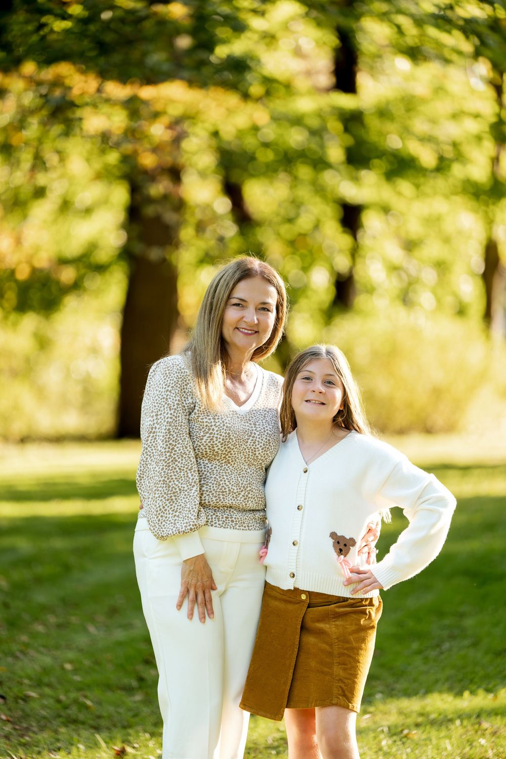 A smiling woman and young girl pose together outdoors in a sunny park with lush green trees in the background.