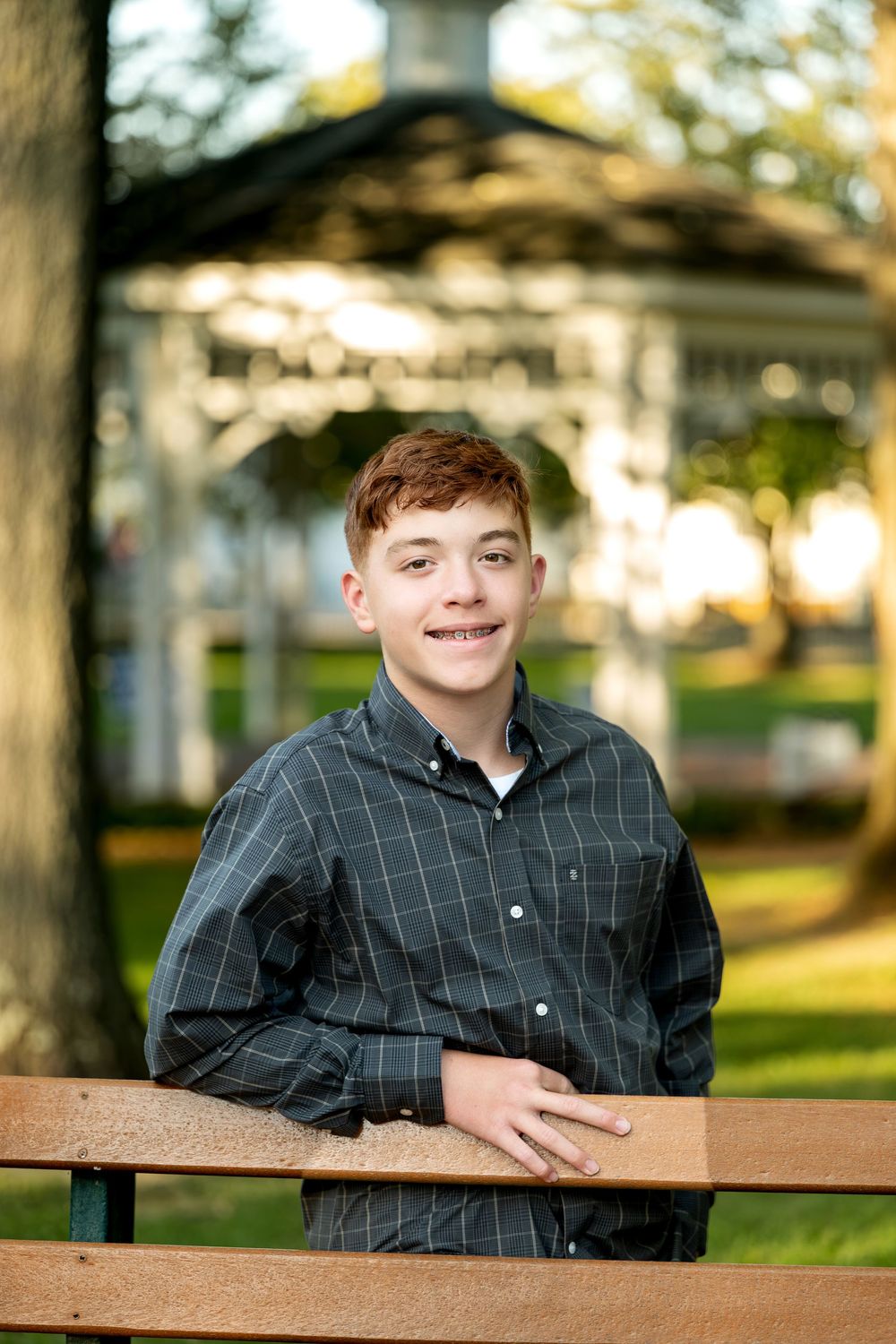 Smiling teenage boy with red hair wearing a plaid shirt, leaning on a park bench with a gazebo in the background.