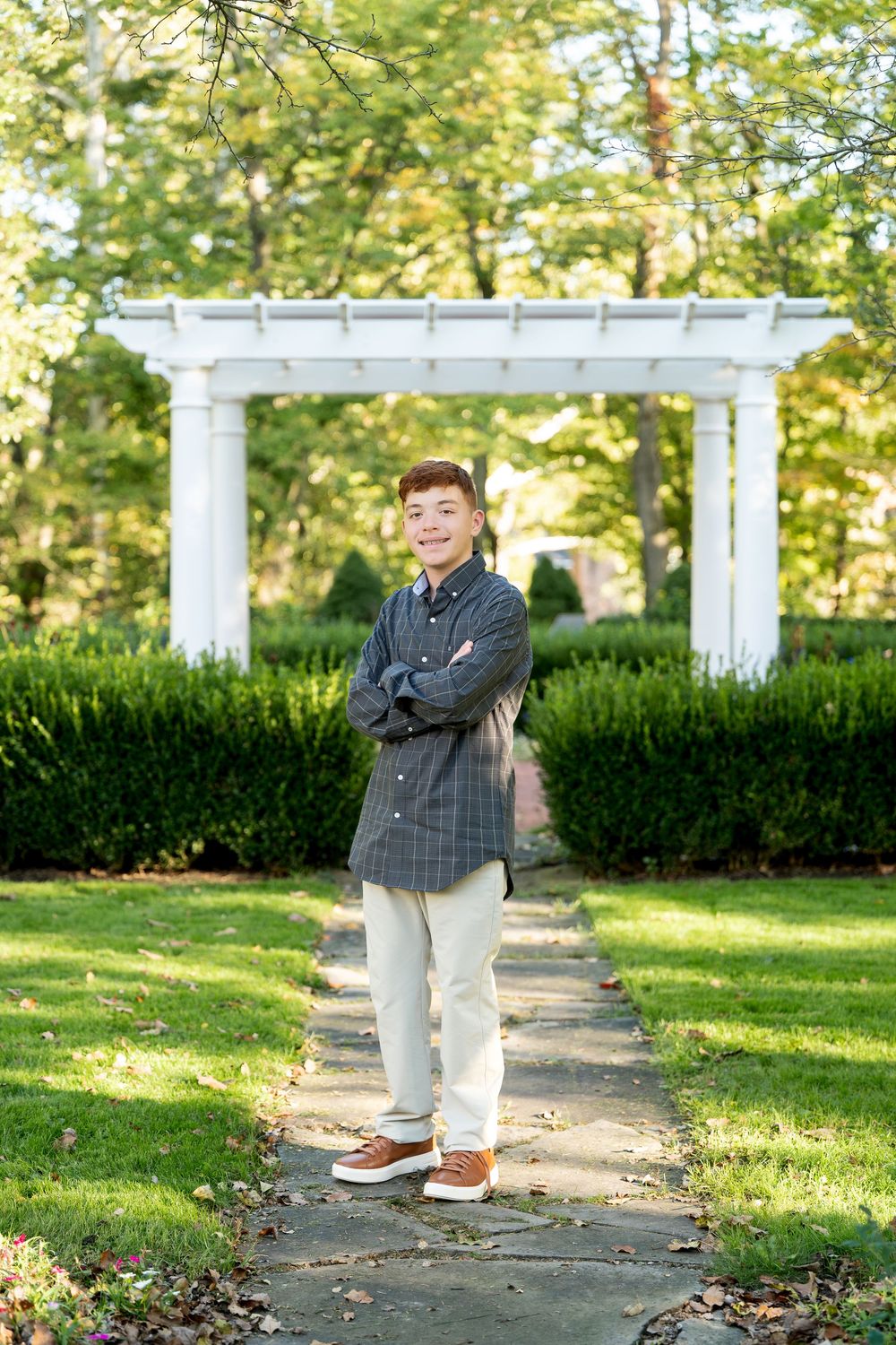 Teen boy in plaid shirt stands arms crossed on garden path with white pergola backdrop.