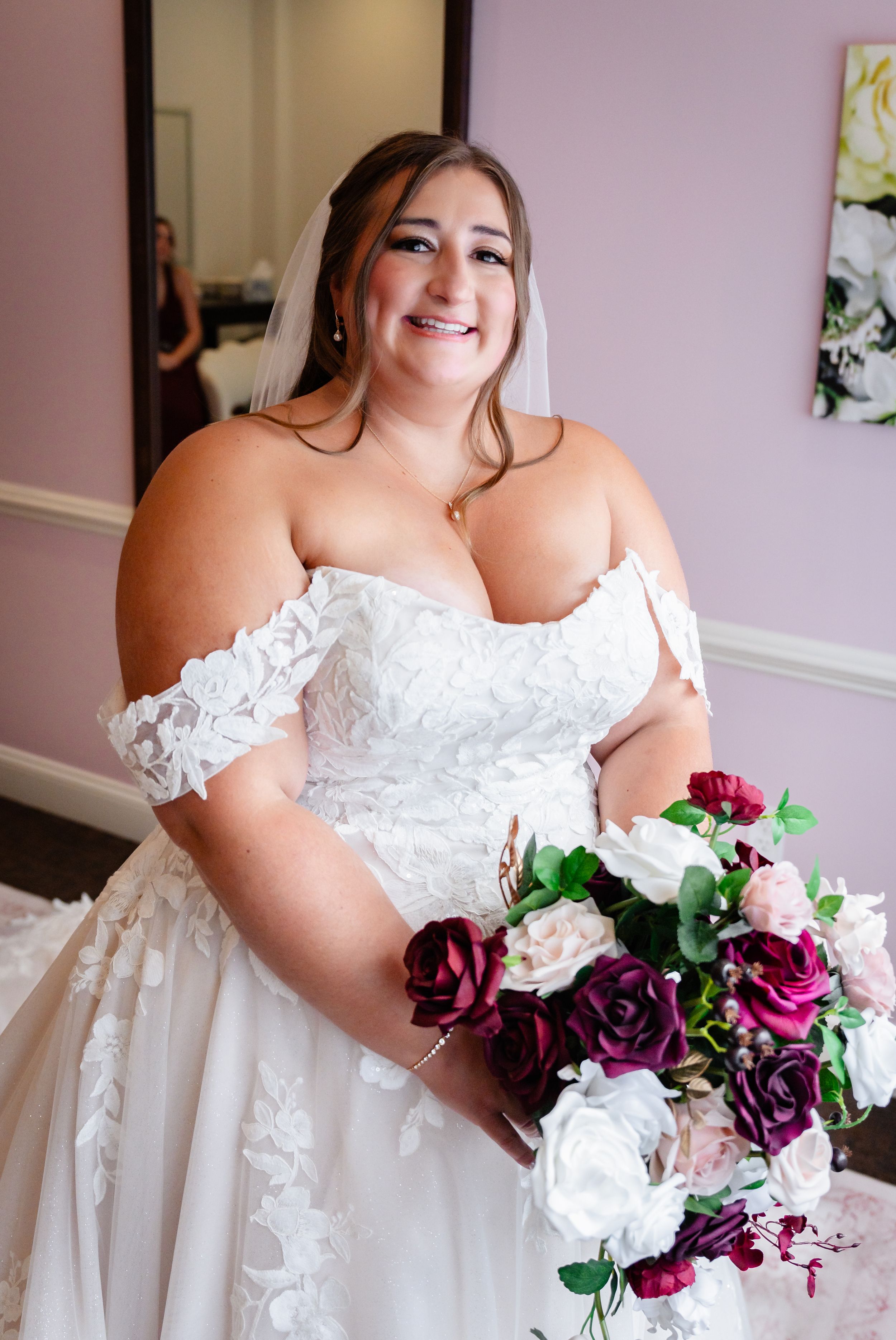 Smiling bride in off-shoulder lace wedding gown holds burgundy and white bouquet.