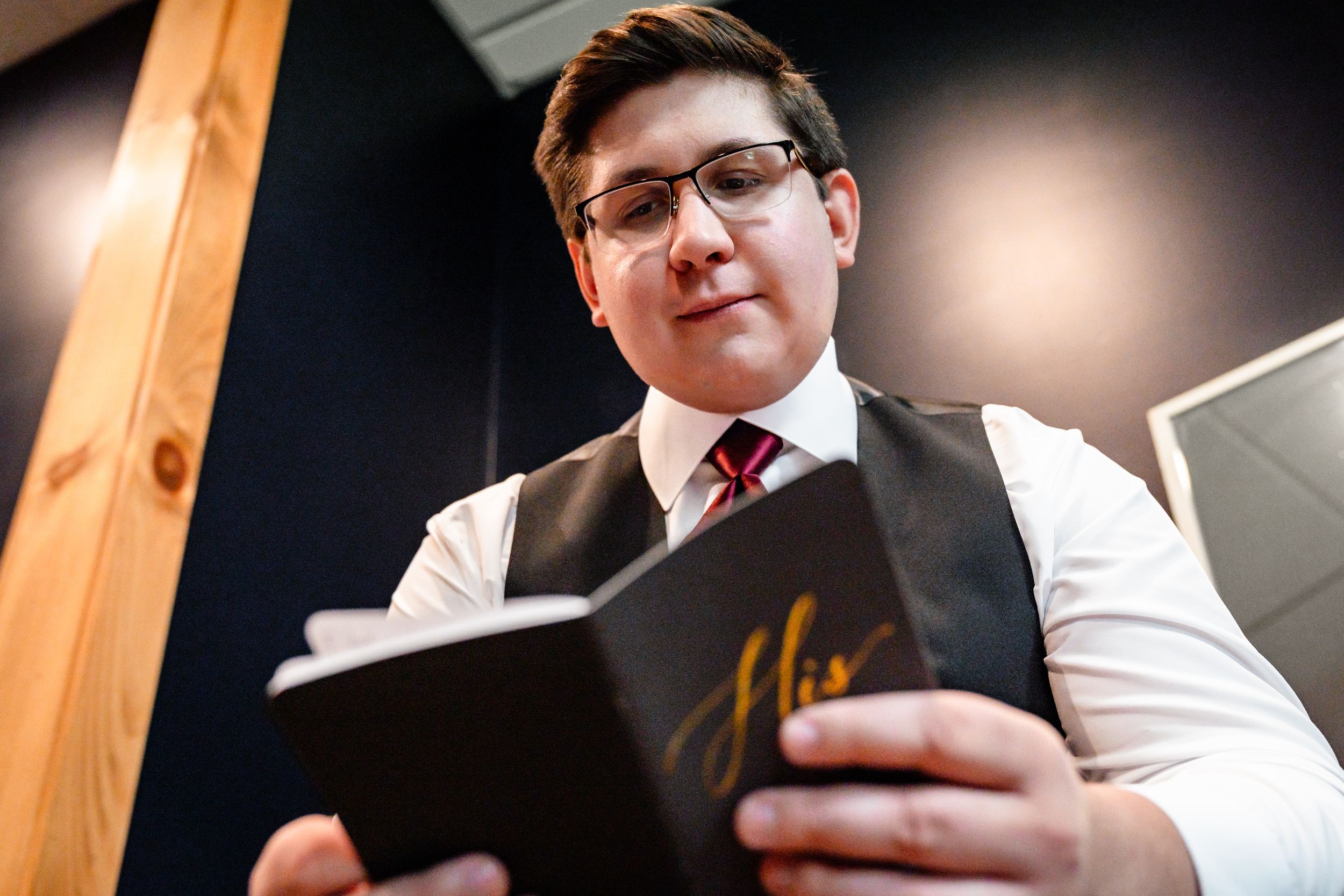 Young man in vest and tie reading a black wedding vow book indoors.