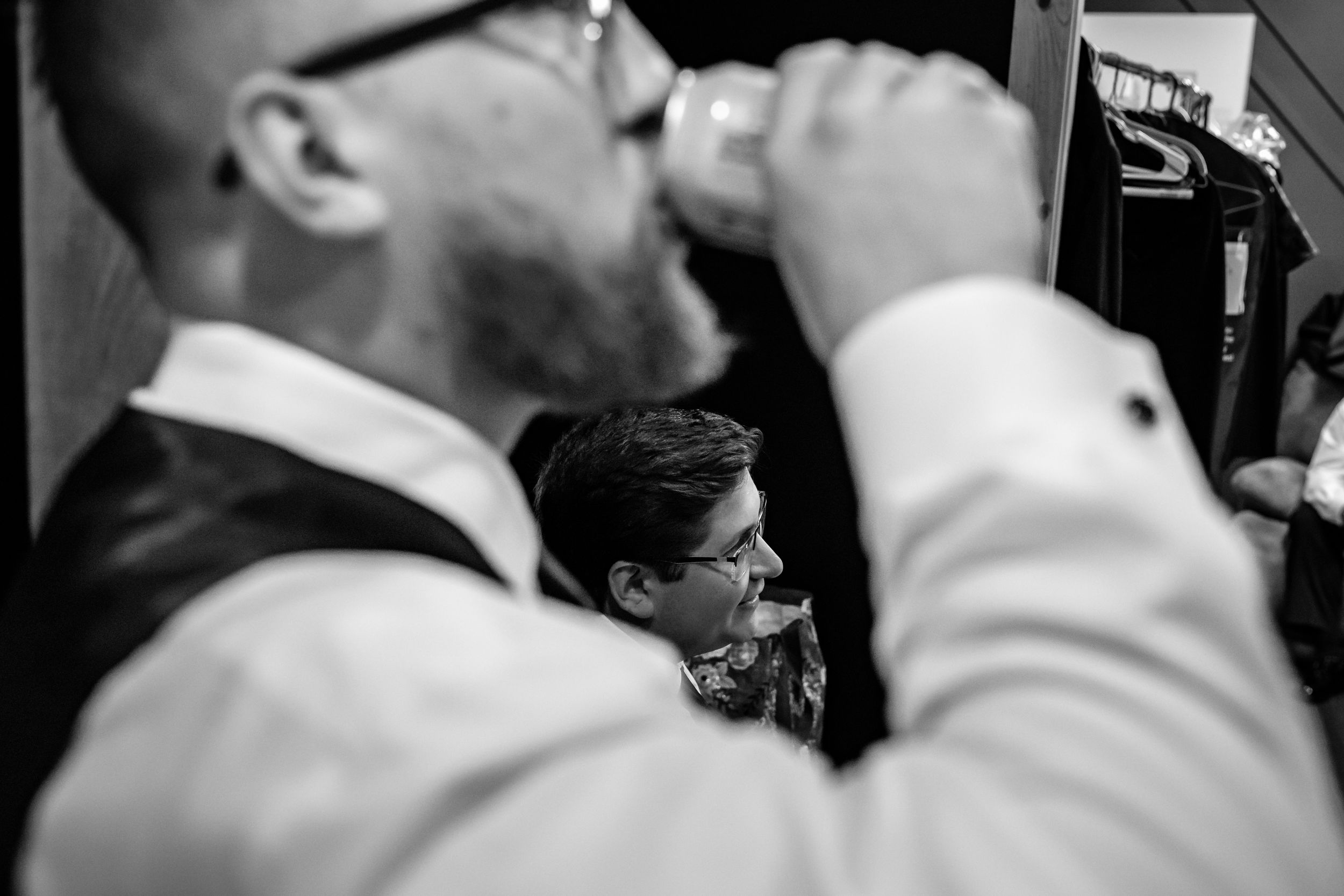 Black and white photo of a groom drinking from a flask while another man adjusts his bow tie at a wedding.