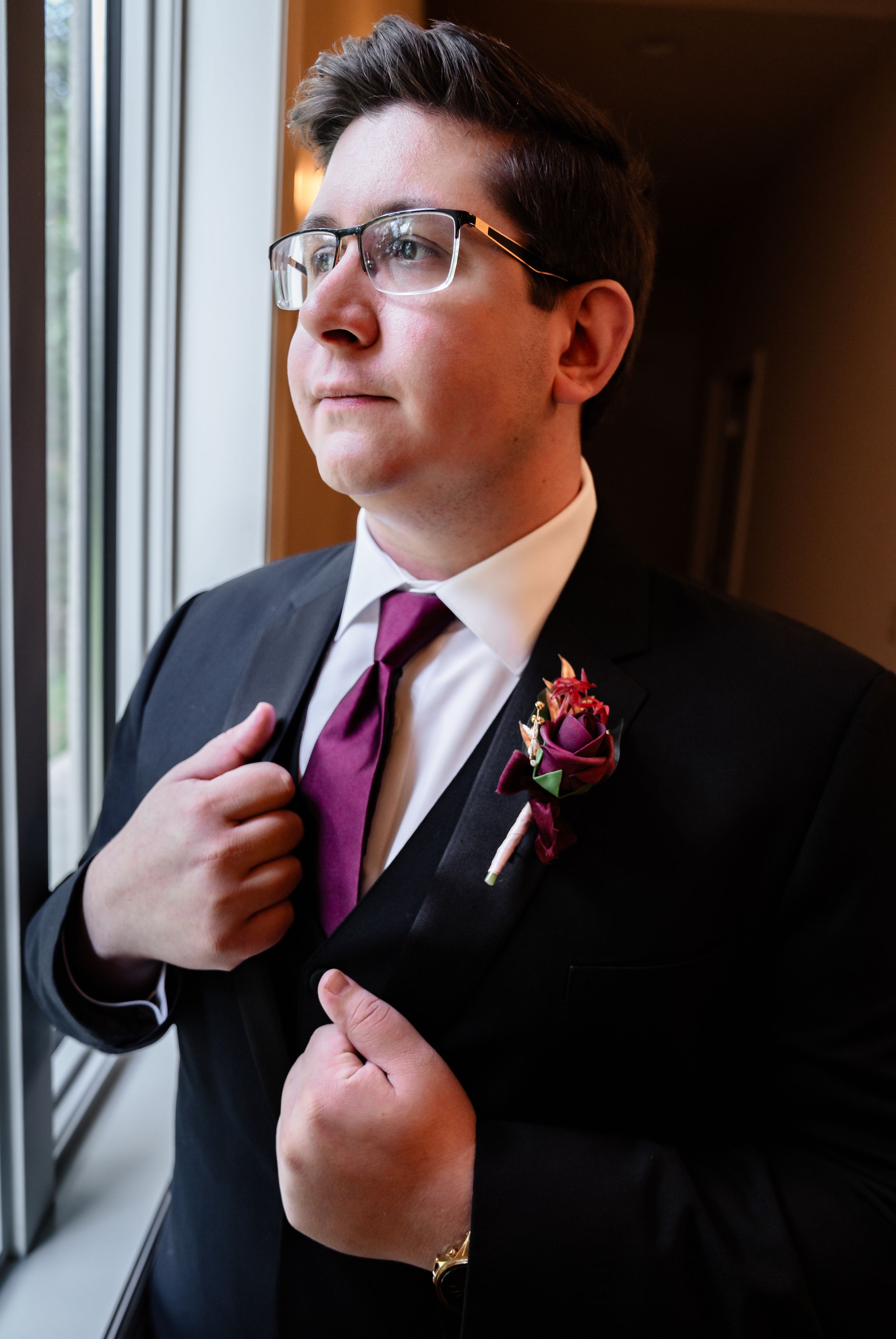 Groom in black suit with purple tie adjusts jacket lapel near window.