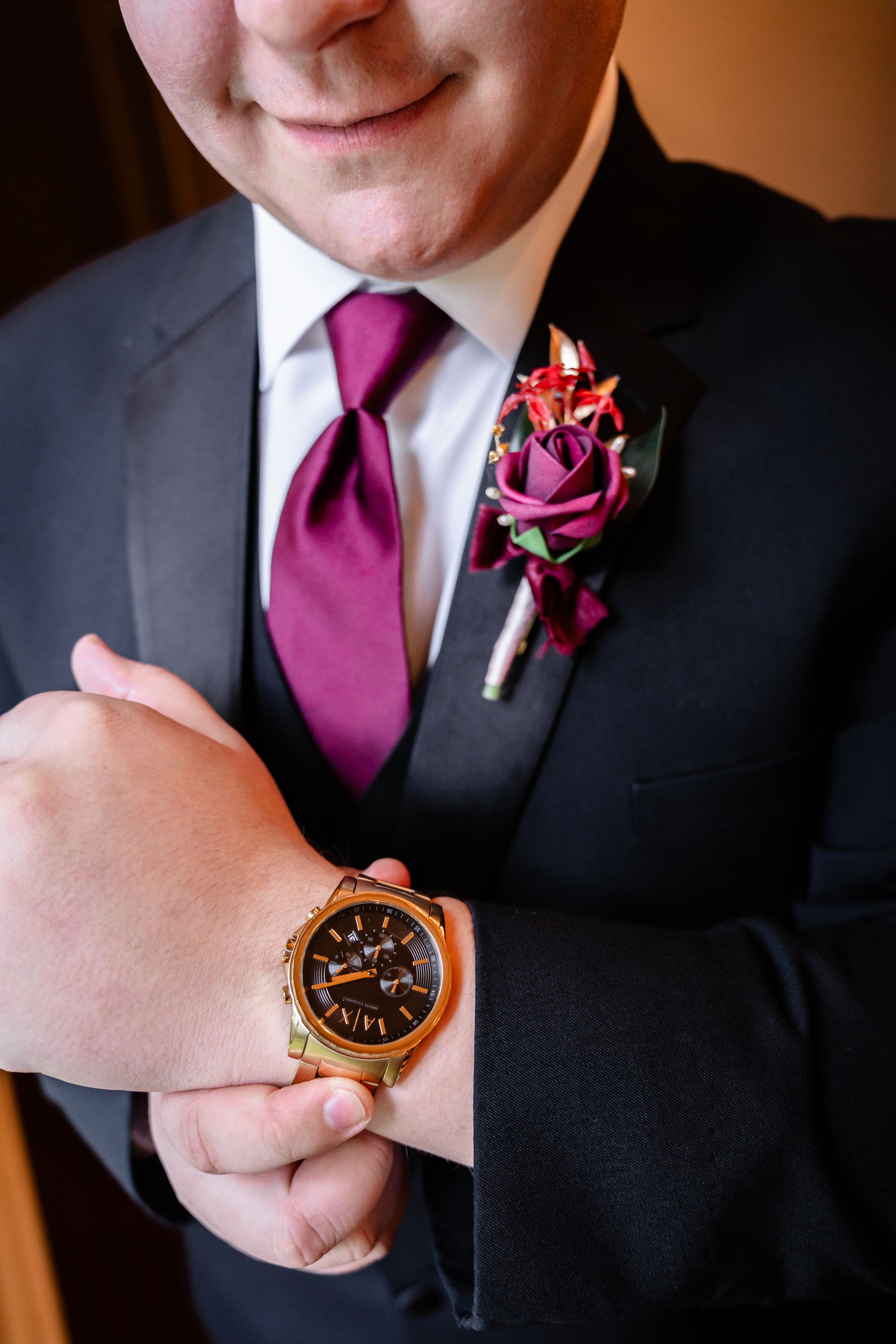 Groom in dark suit adjusting gold watch, wearing burgundy tie and floral boutonniere.
