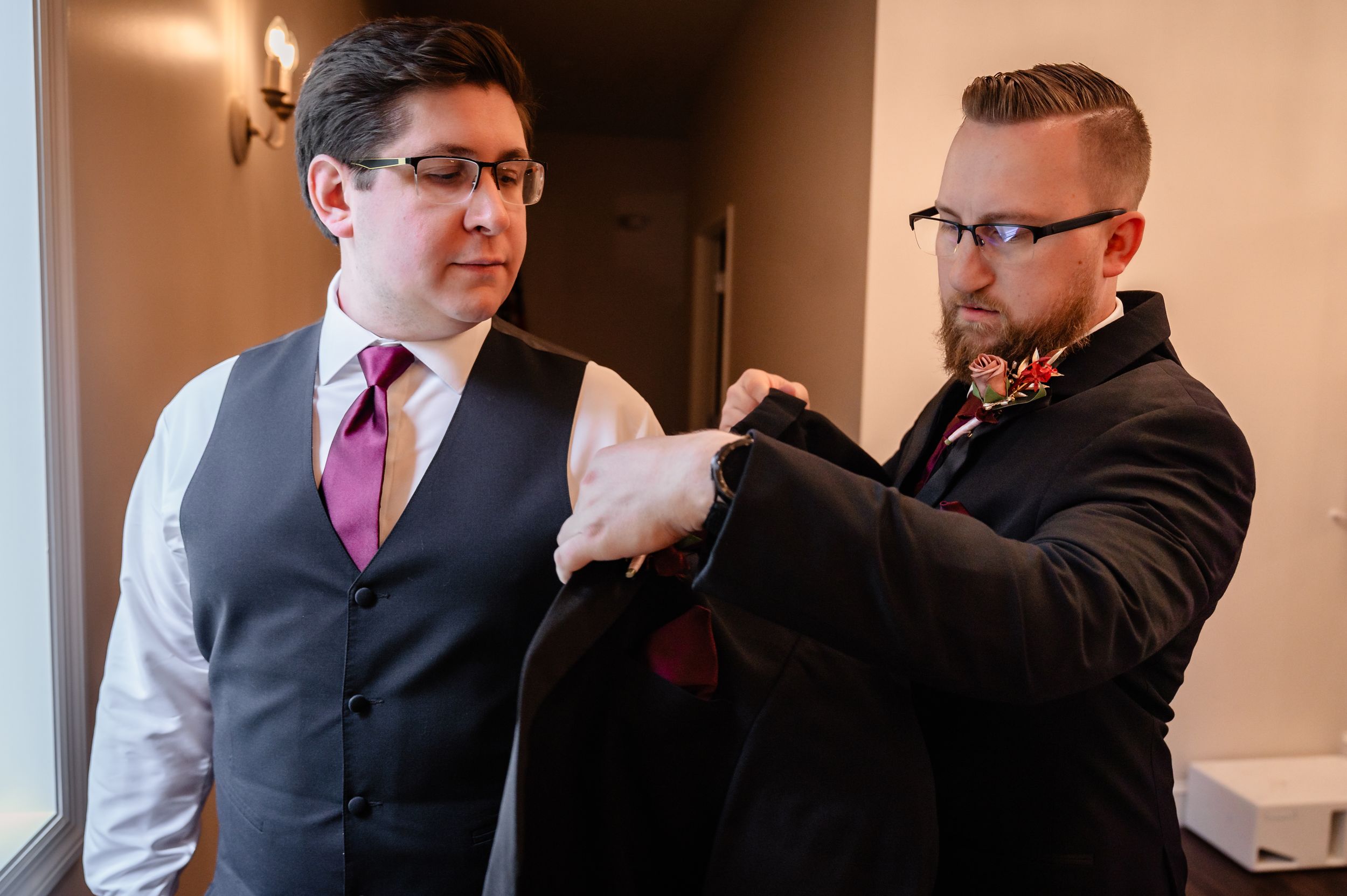 Groomsman helping groom adjust suit jacket before wedding ceremony indoors.