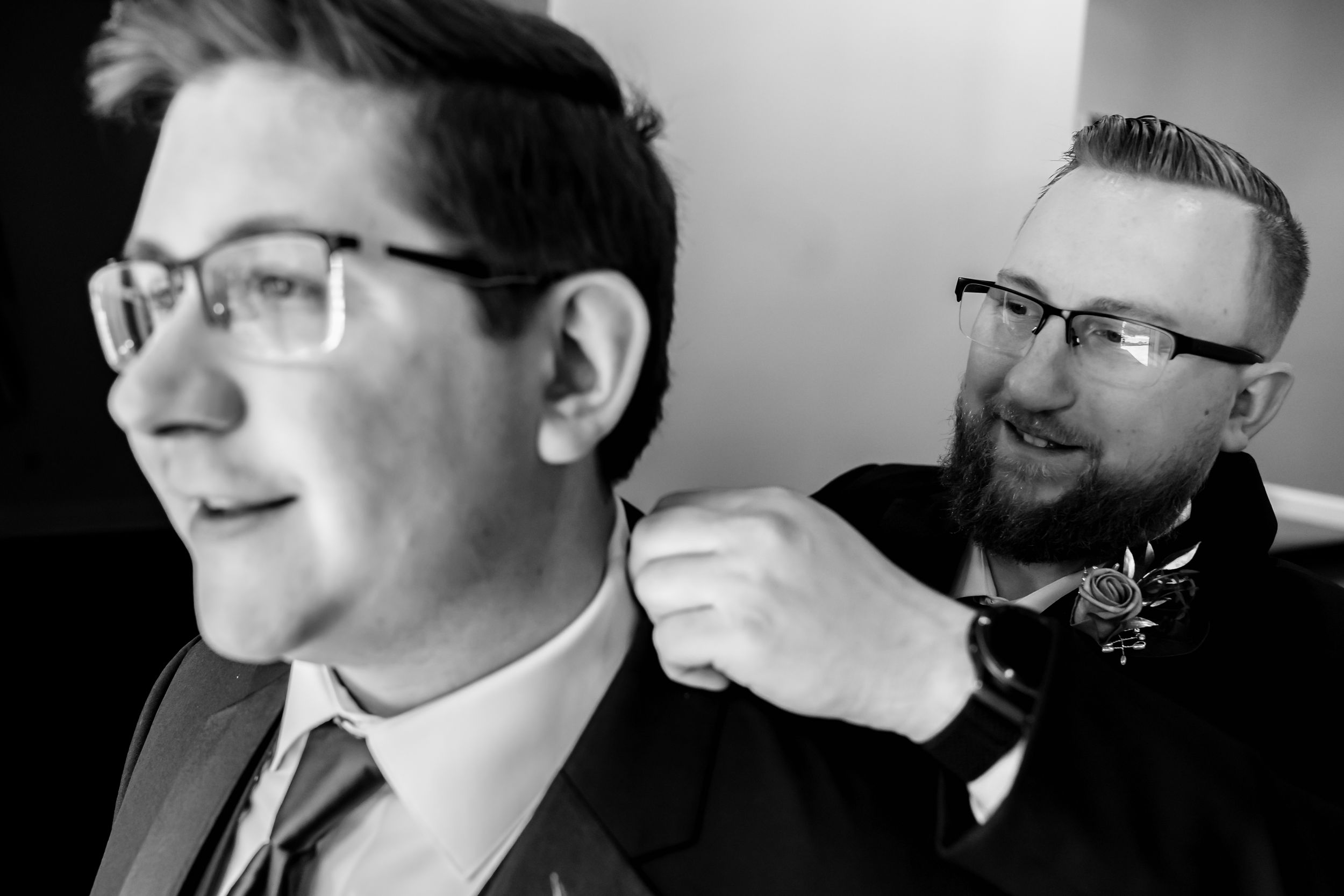 Man in glasses adjusting bow tie of younger man in suit, black and white wedding preparation photo.