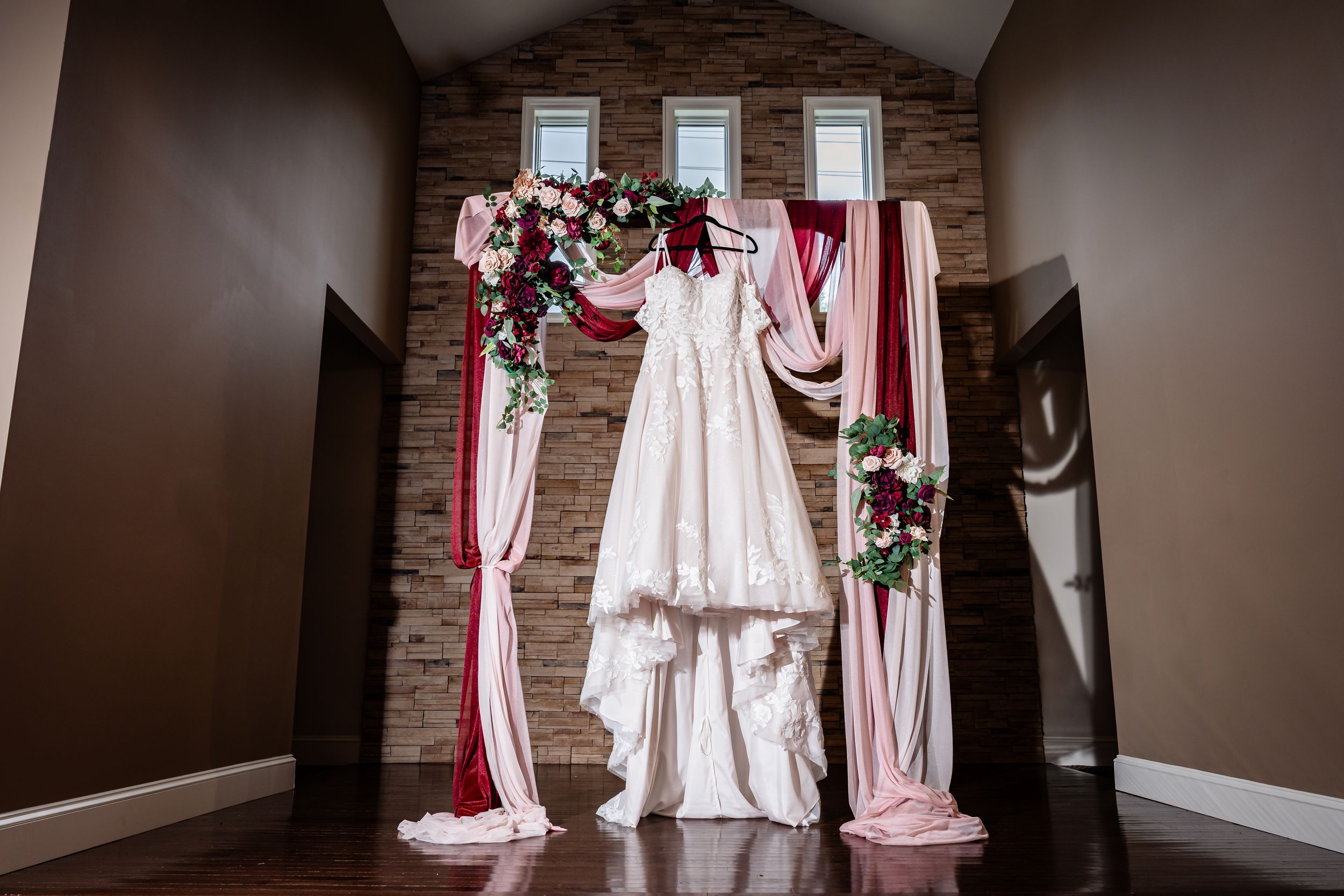 Elegant wedding dress displayed on a floral arch with pink and burgundy draping against a stone wall.