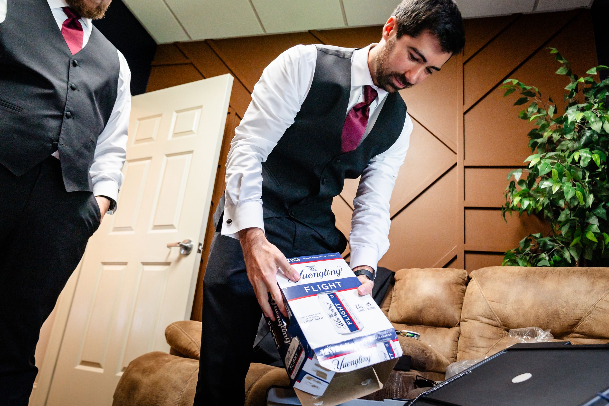 Groomsman in vest opens wedding-related box on couch before ceremony.