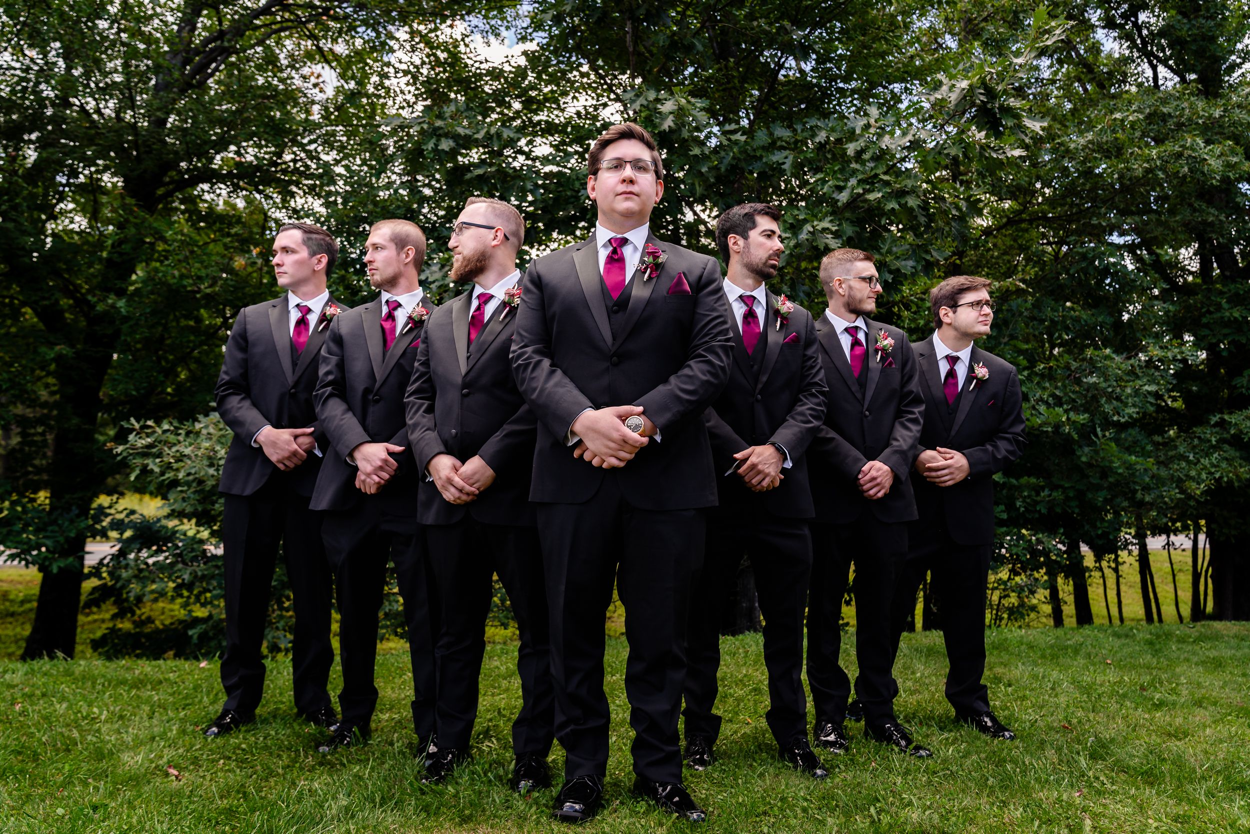 Groom stands confidently with groomsmen in matching gray suits on green lawn.