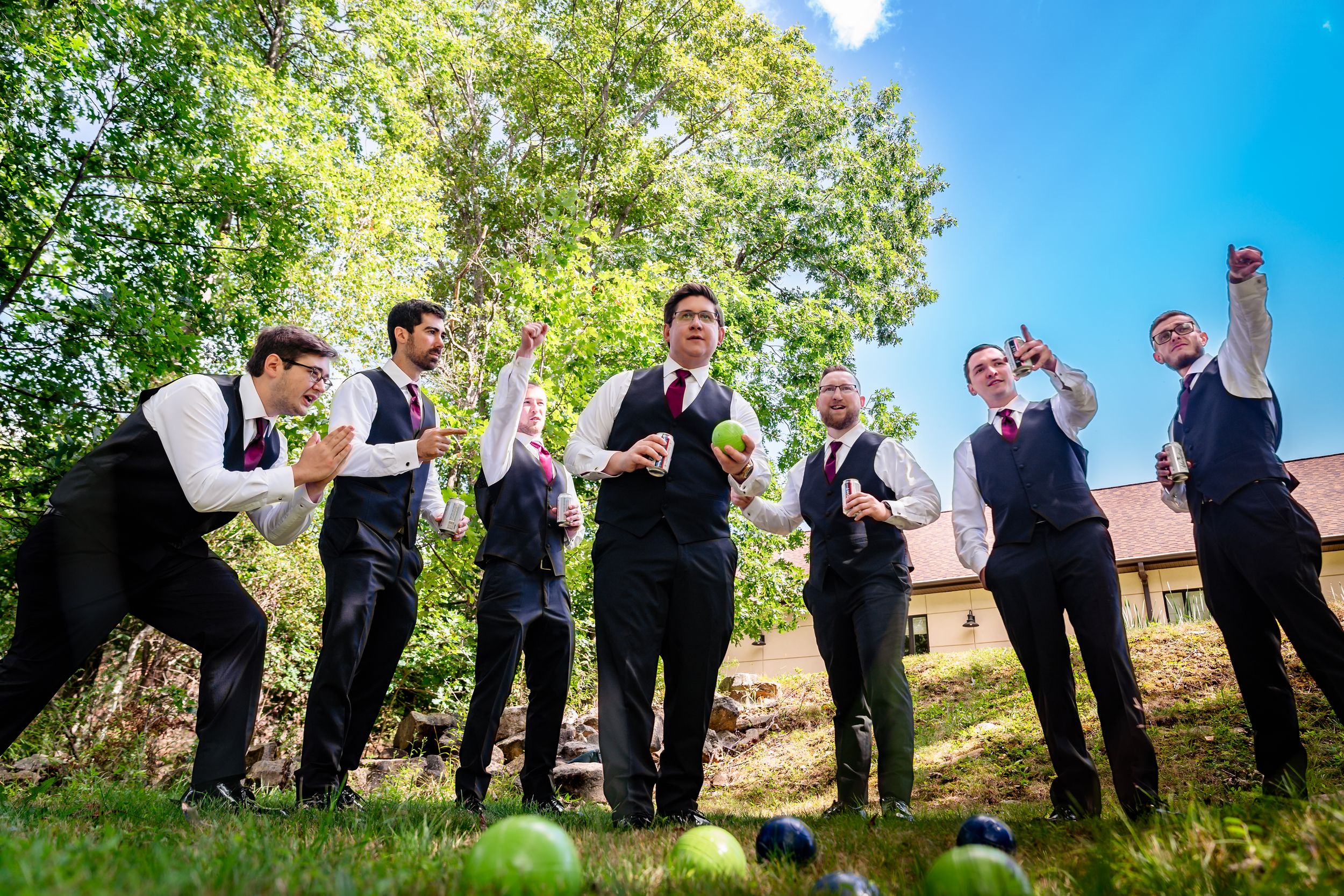 Groomsmen in black suits and burgundy ties celebrating outdoors on a sunny day at a wedding.