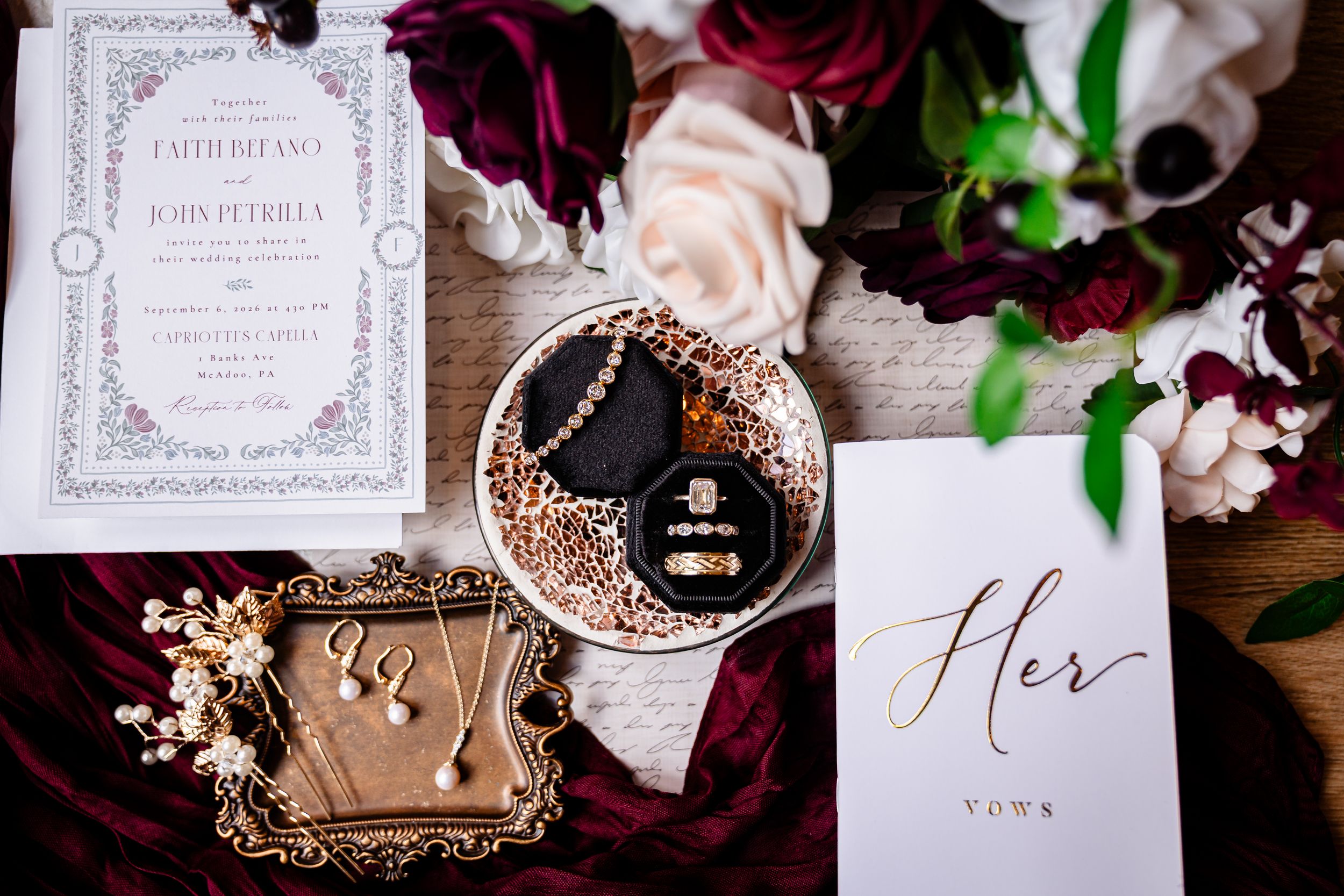 Elegant wedding flat lay with floral invitations, gold jewelry box, rings, earrings, and Her Vows card with burgundy roses.