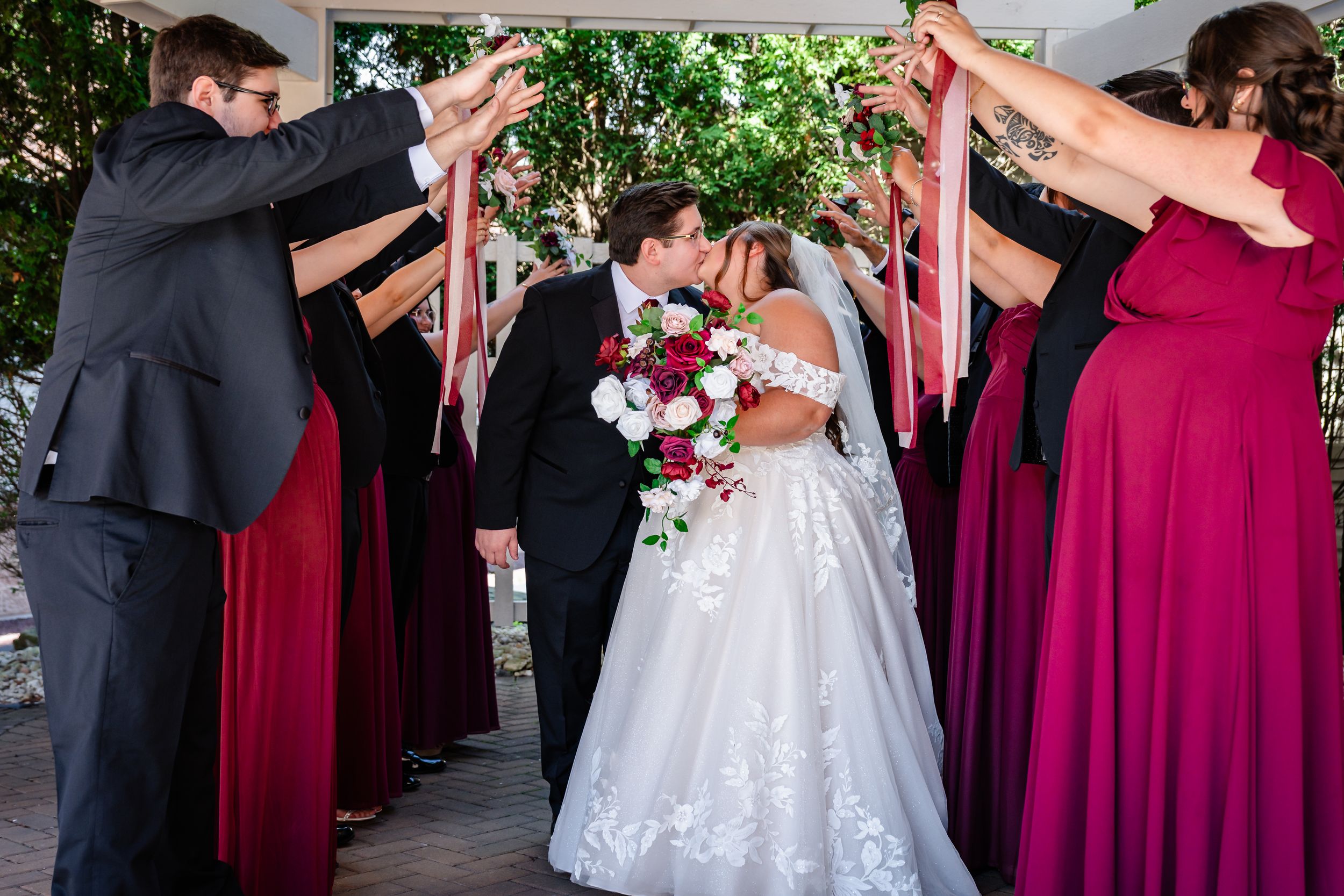 Bride and groom share a kiss under an arch of raised hands formed by bridesmaids in red gowns and groomsmen.