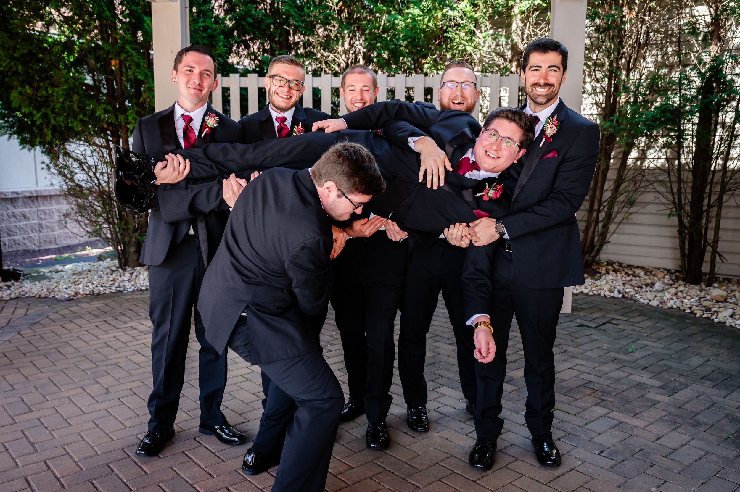 Groomsmen in black suits playfully posing outdoors with an elderly couple at a wedding celebration.