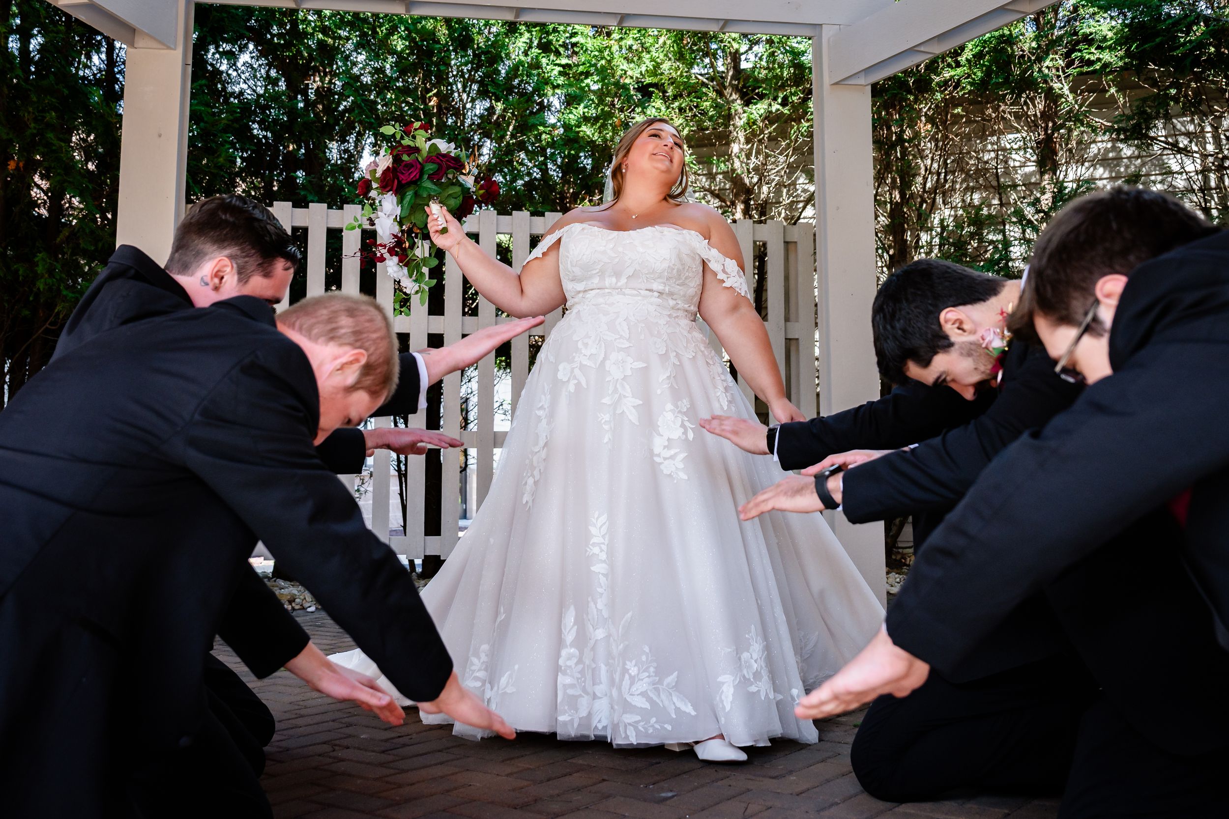 Bride in white gown posing with groomsmen bowing at outdoor wedding venue.