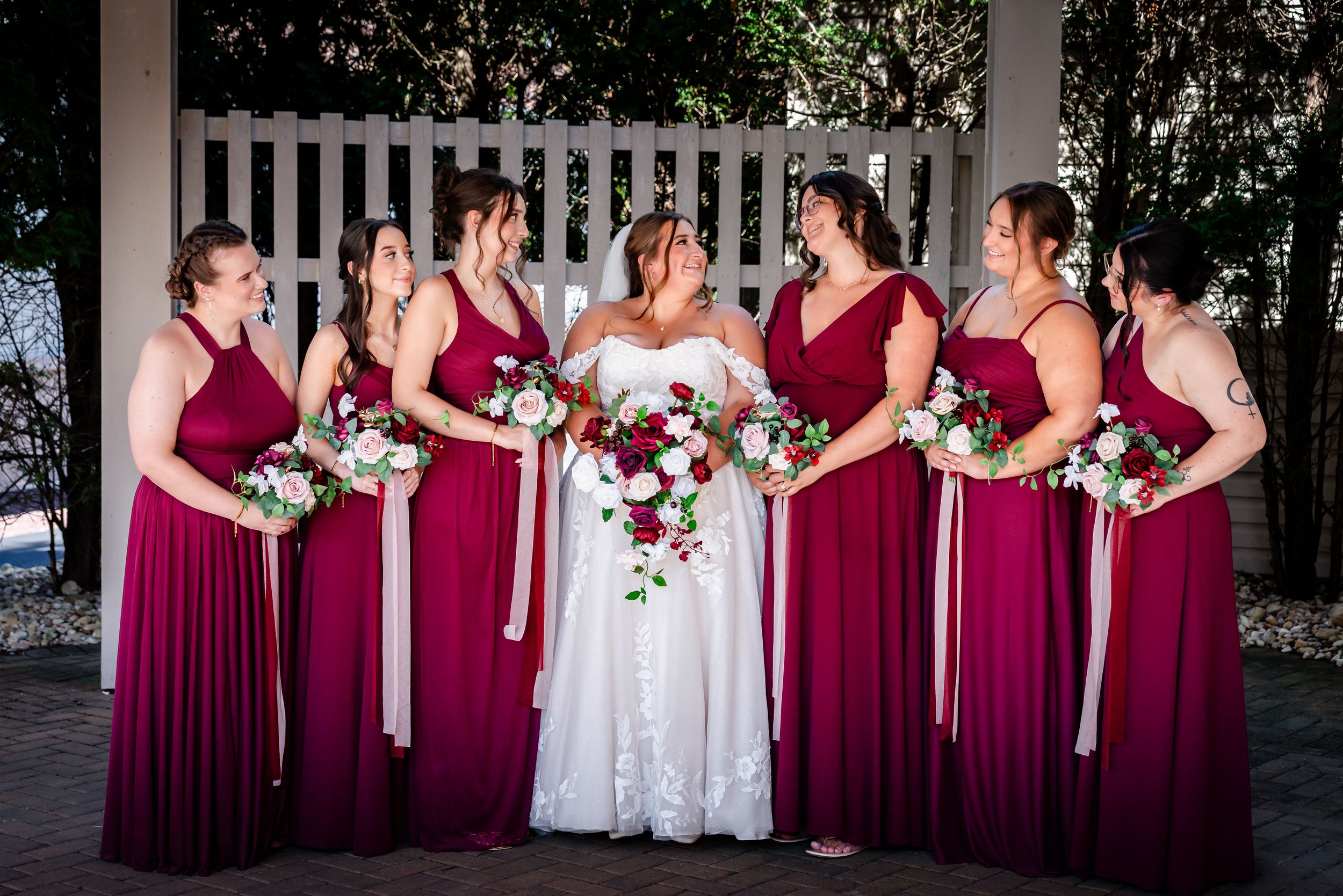 Bride in white gown surrounded by six bridesmaids in burgundy dresses holding floral bouquets outdoors.