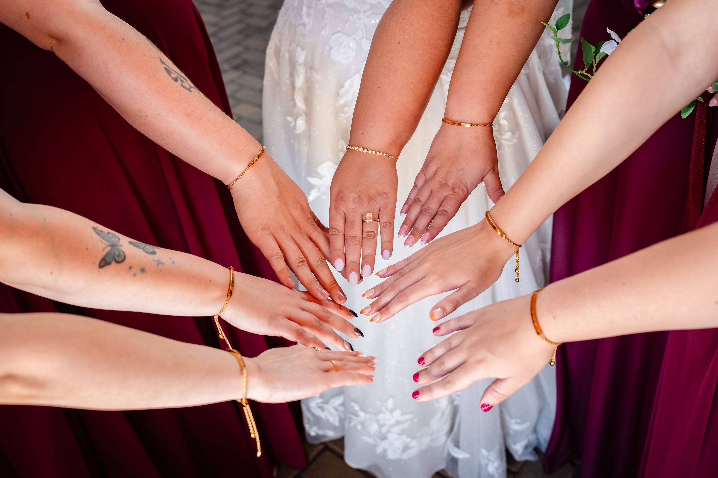 Bridesmaids in burgundy dresses placing hands on brides white gown showing gold bracelets and manicured nails.