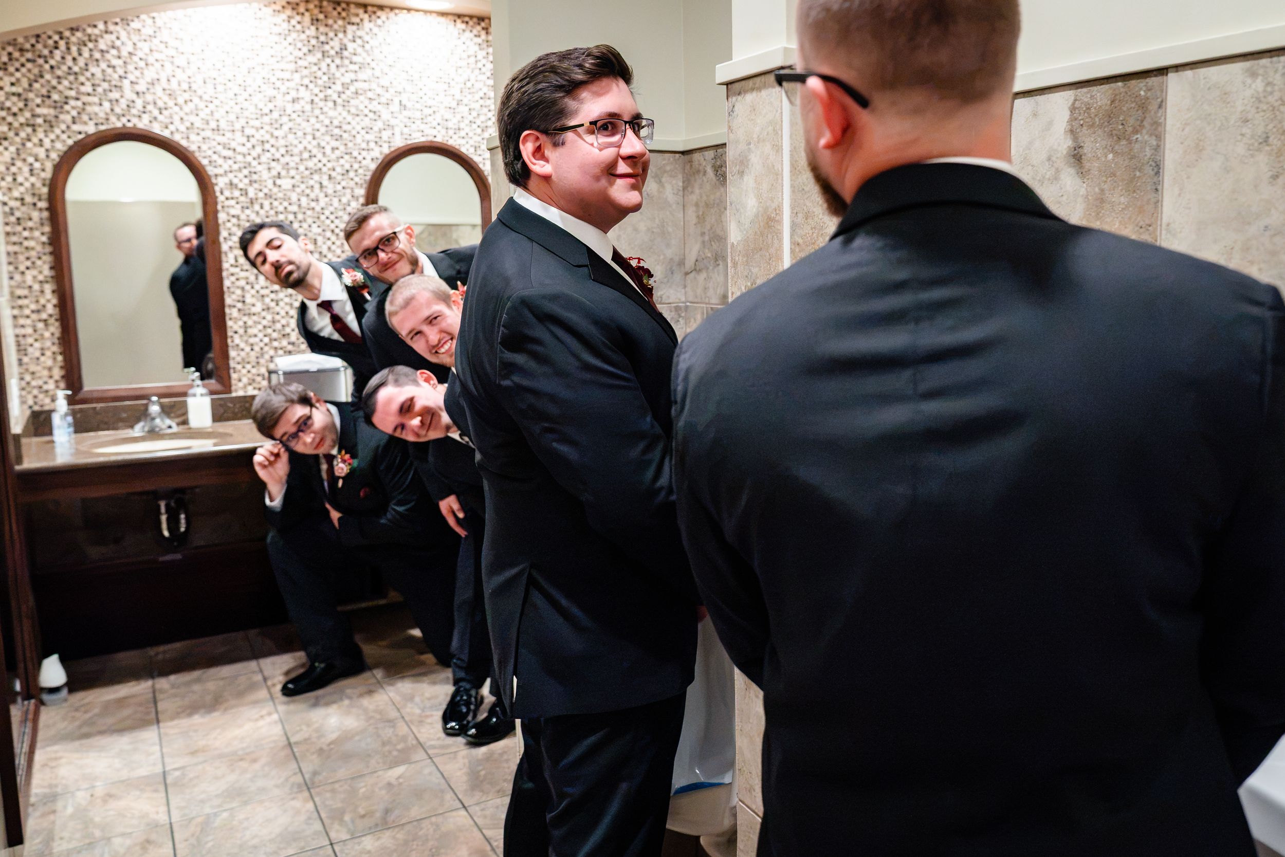 Groom laughs with groomsmen in tiled bathroom while getting ready.