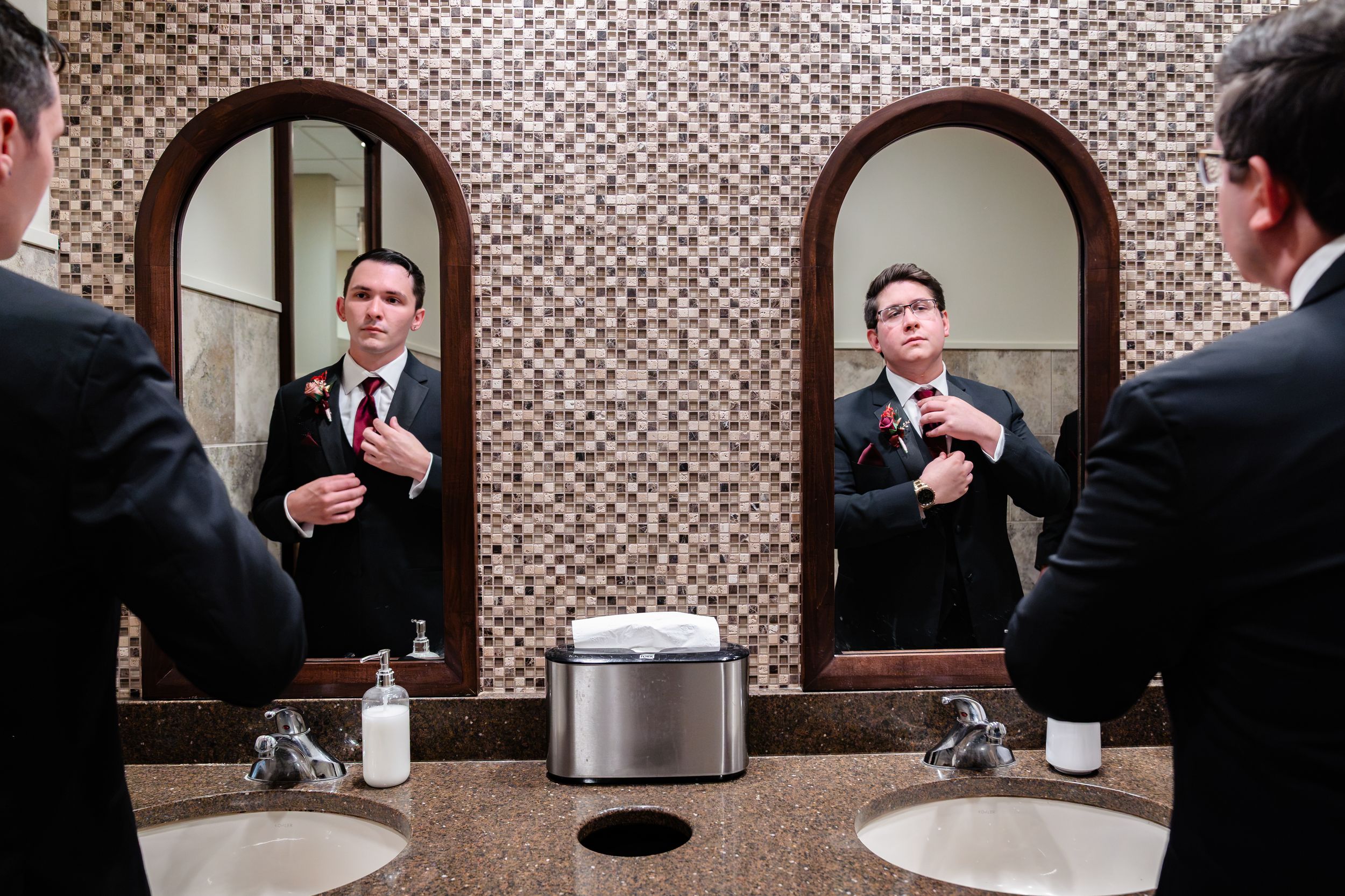 Groom adjusts boutonniere while reflected in ornate bathroom mirrors.