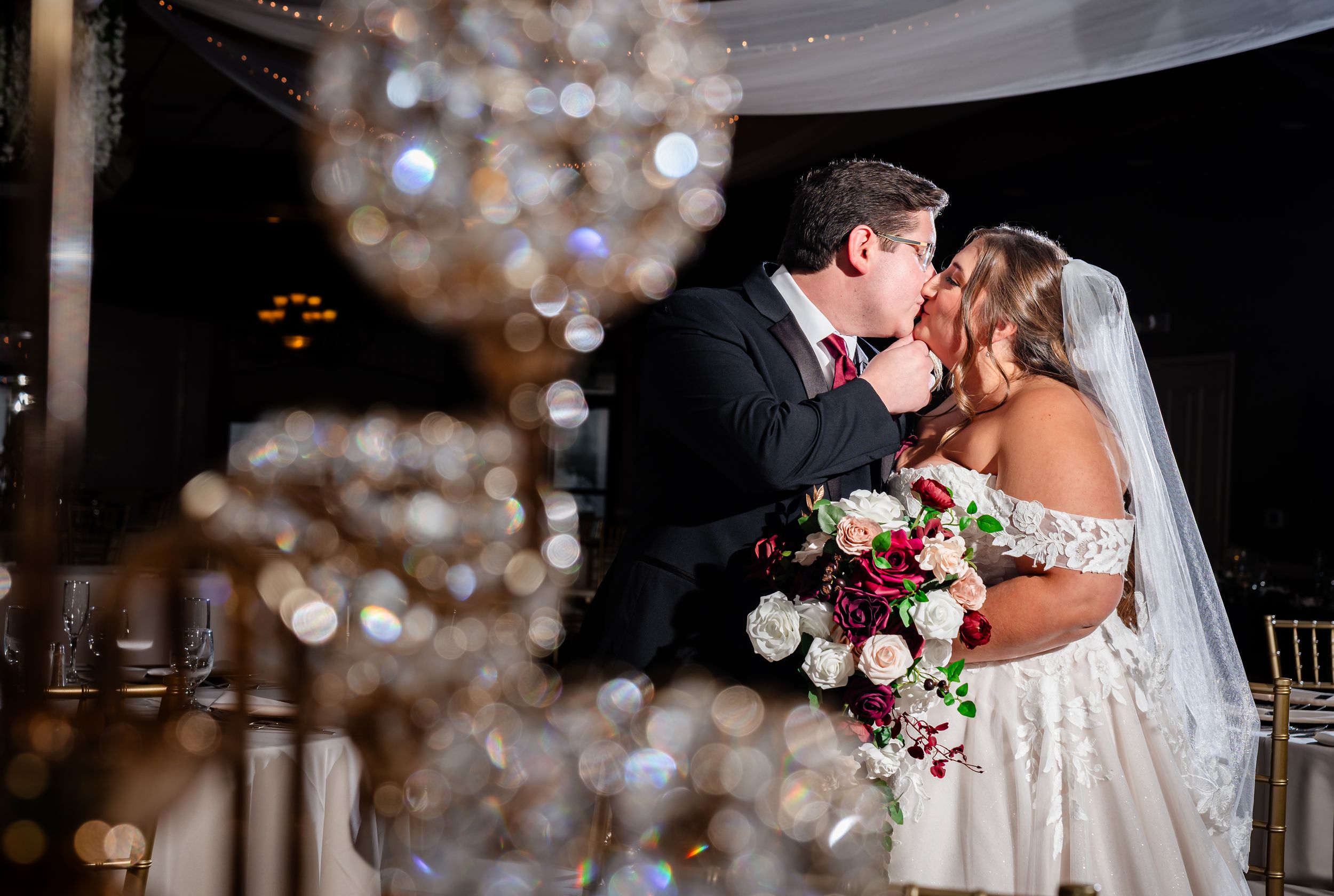 Bride and groom share first kiss framed by hanging crystal chandeliers at wedding reception.