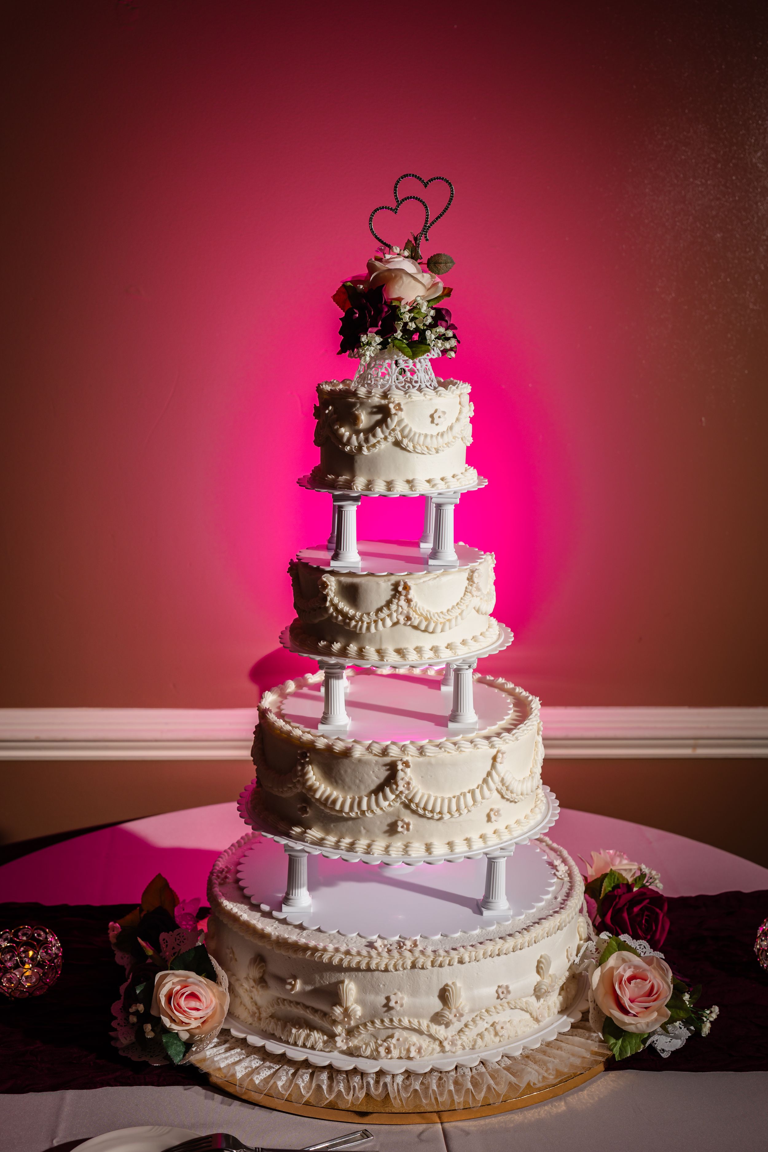Elegant four-tier white wedding cake with intricate piping, floral decorations, and heart topper against a pink wall.