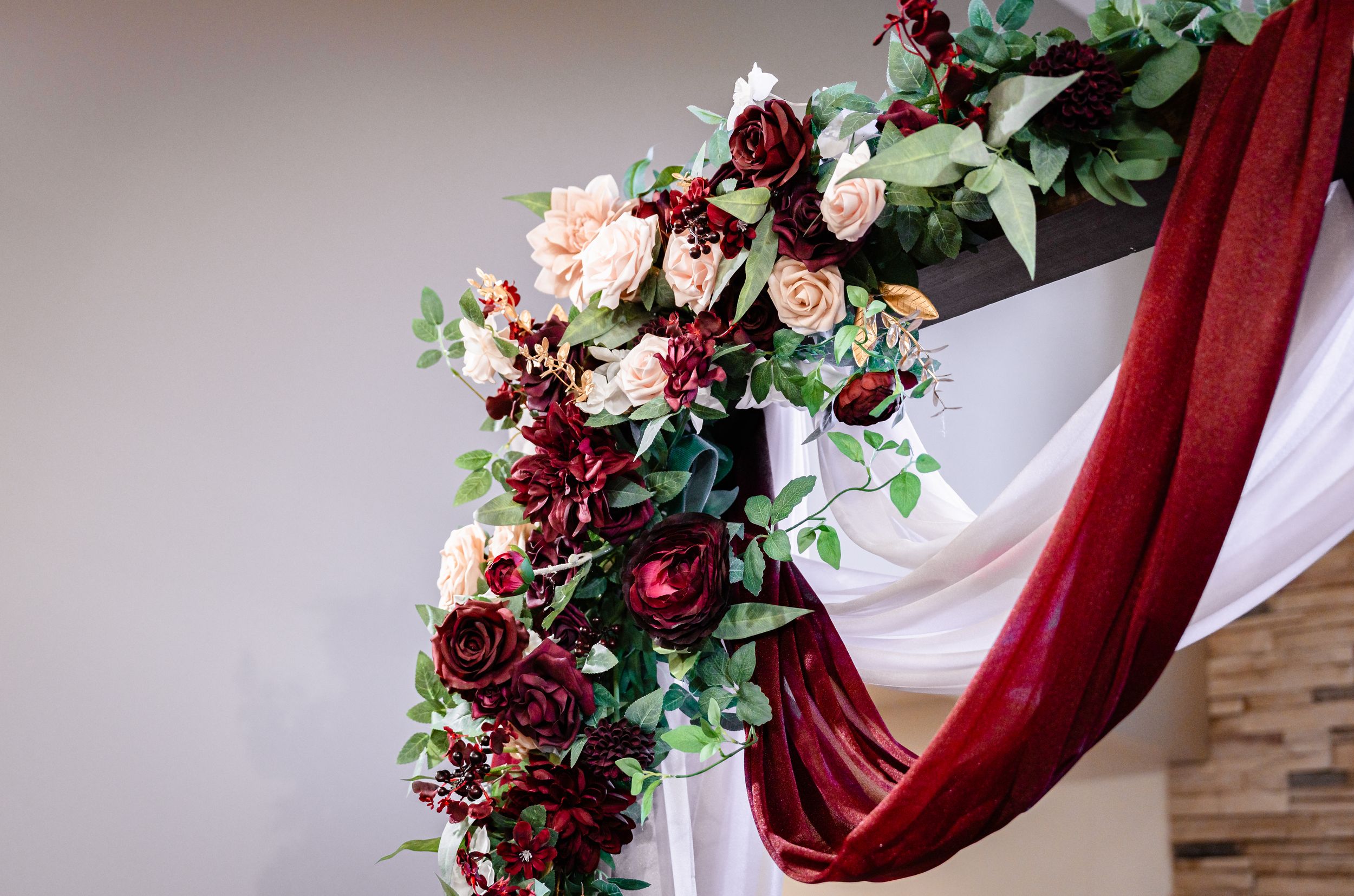 Elegant wedding arch decorated with burgundy and blush roses, greenery, and flowing red and white draping.