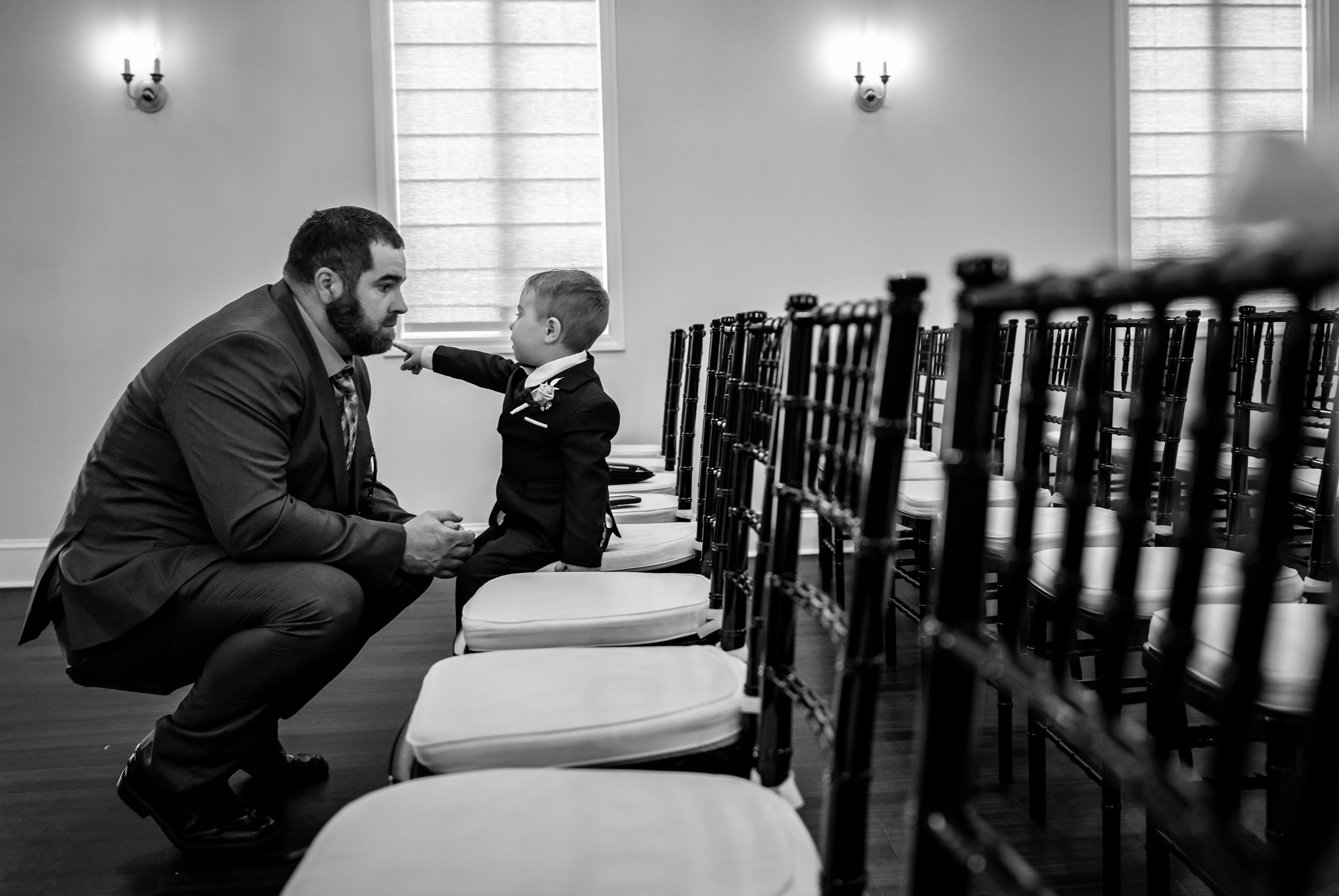 A man crouches down to interact with a young boy in a suit between ceremony chairs in black and white.